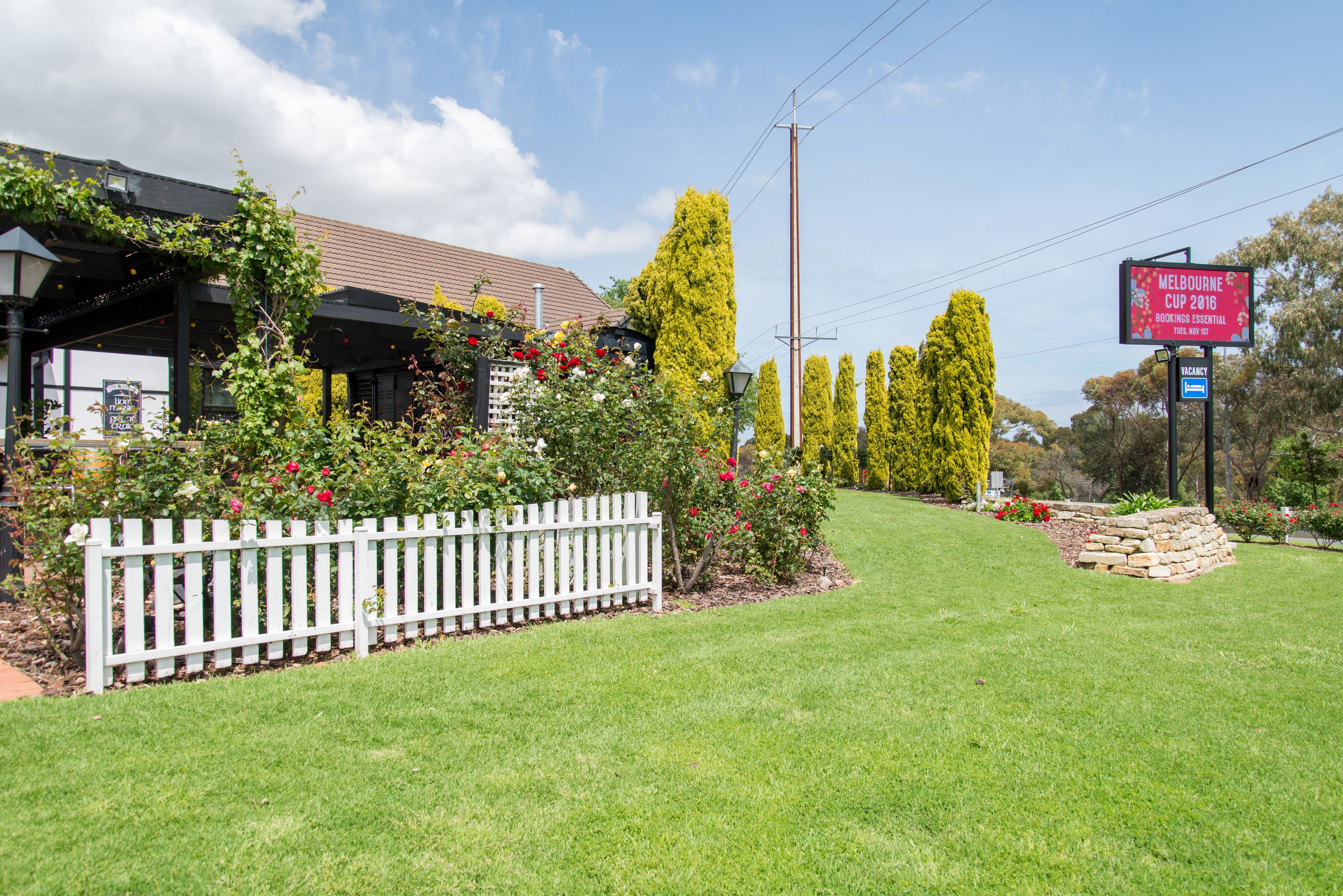 Un hotel con una valla blanca de piquetes, un espacio al aire libre cubierto con plantas y un cartel anunciando la Copa de Melbourne 2016.