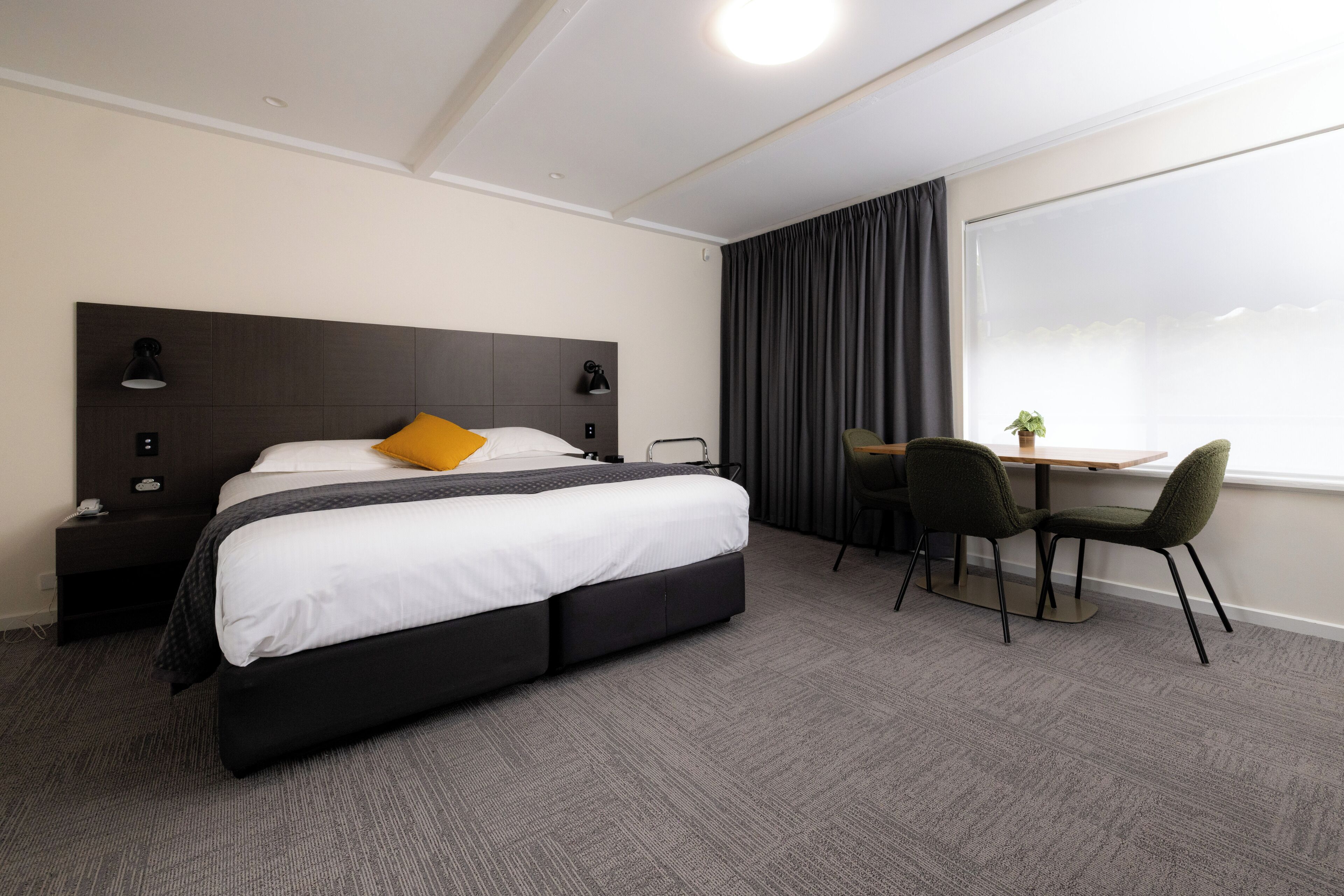 Habitación de hotel con una cama grande, un escritorio con dos sillas, una ventana con cortinas y una mesita de noche con lámpara.