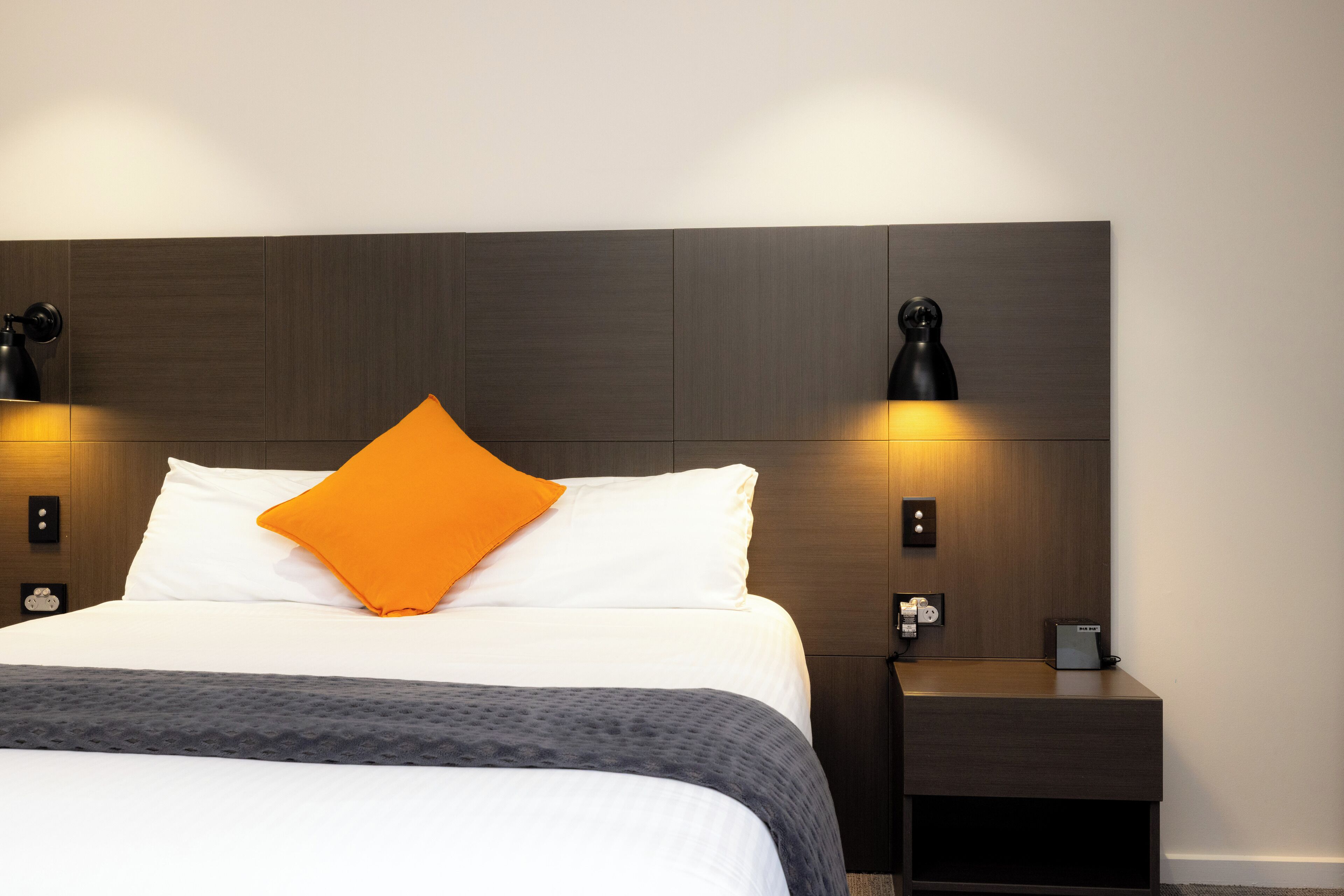 Una cama bien hecha con sábanas blancas y un almohadón naranja, flanqueada por dos apliques negros en la pared.