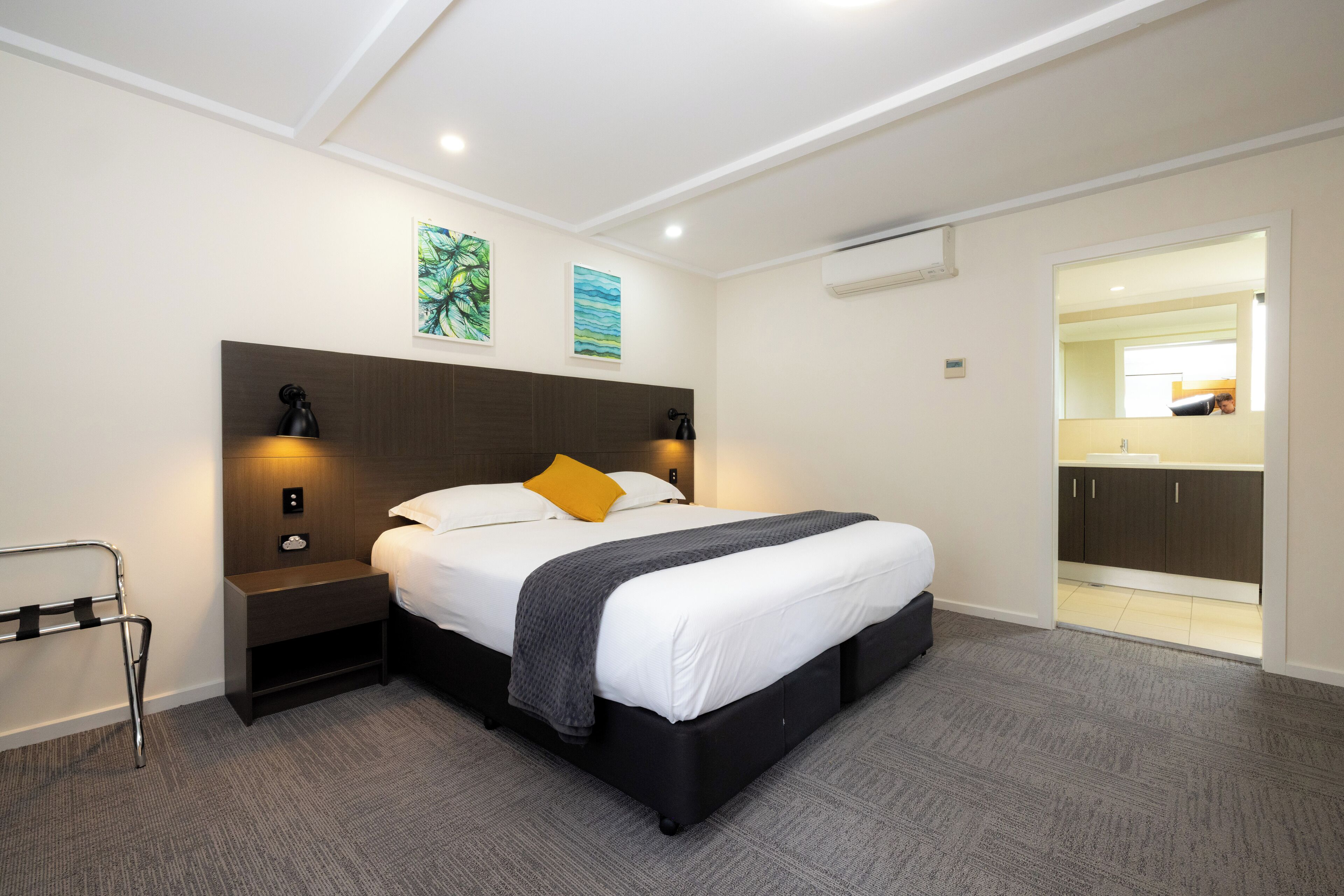 Una habitación de hotel moderna con una cama grande, mesitas de noche, una mesita auxiliar, una silla de ruedas y un baño visible a través de una puerta abierta.