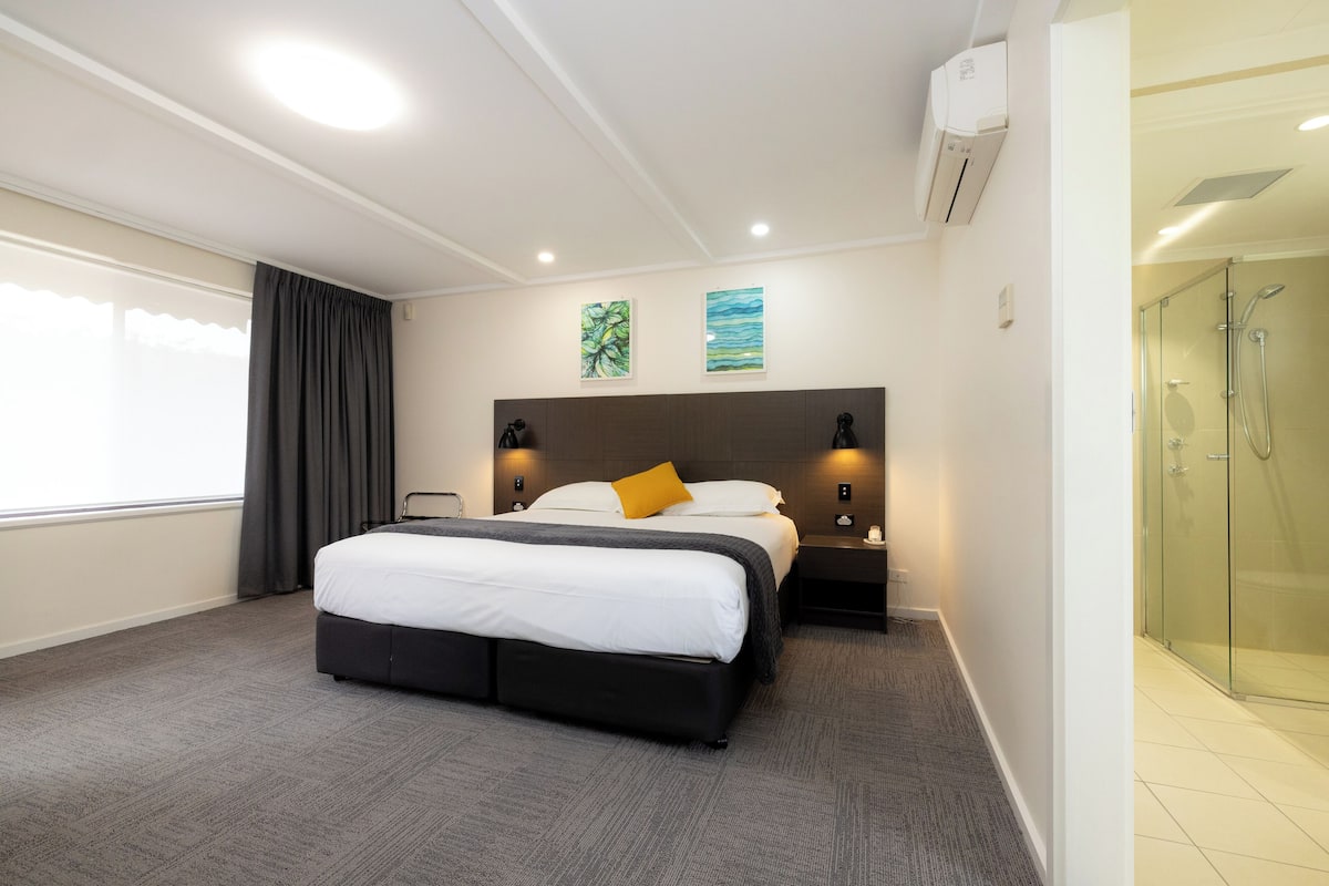 A hotel room with a large bed, a bedside table, a nightstand, a glass shower enclosure, and two wall-mounted artworks.