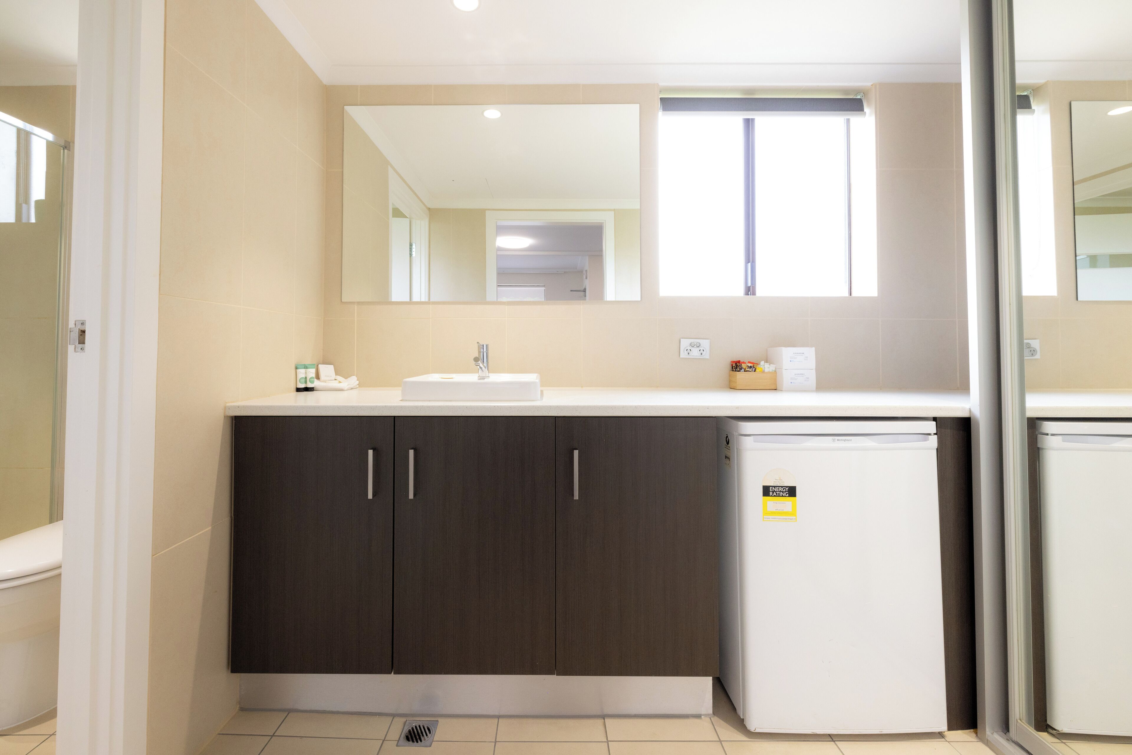 A modern bathroom with a white sink, dark wooden cabinets, a small white refrigerator, and a mirror above the sink.