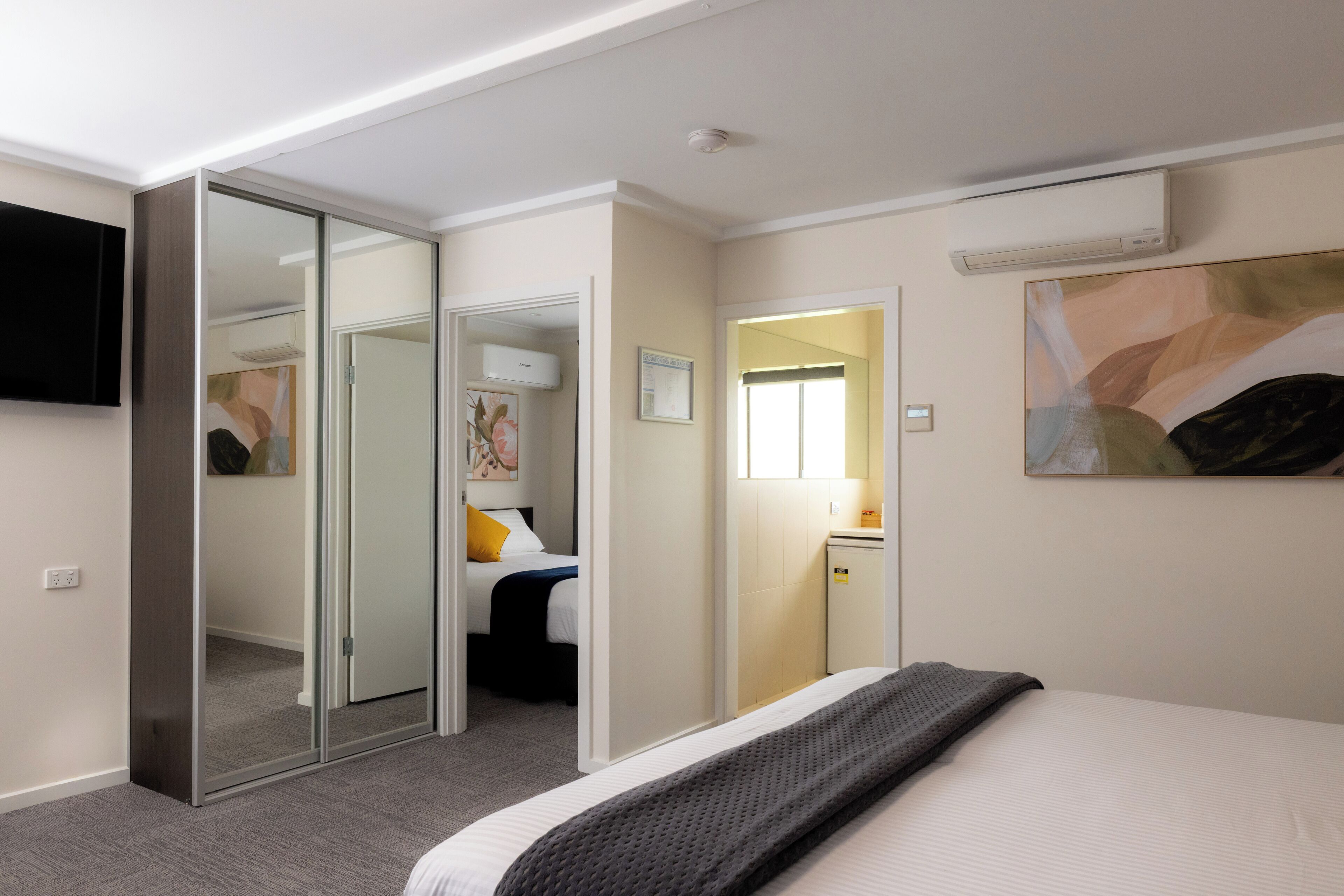 Et hotelværelse med en seng, et fjernsyn ophængt på væggen, en skydelågeskab og et lille skrivebord med en lampe.