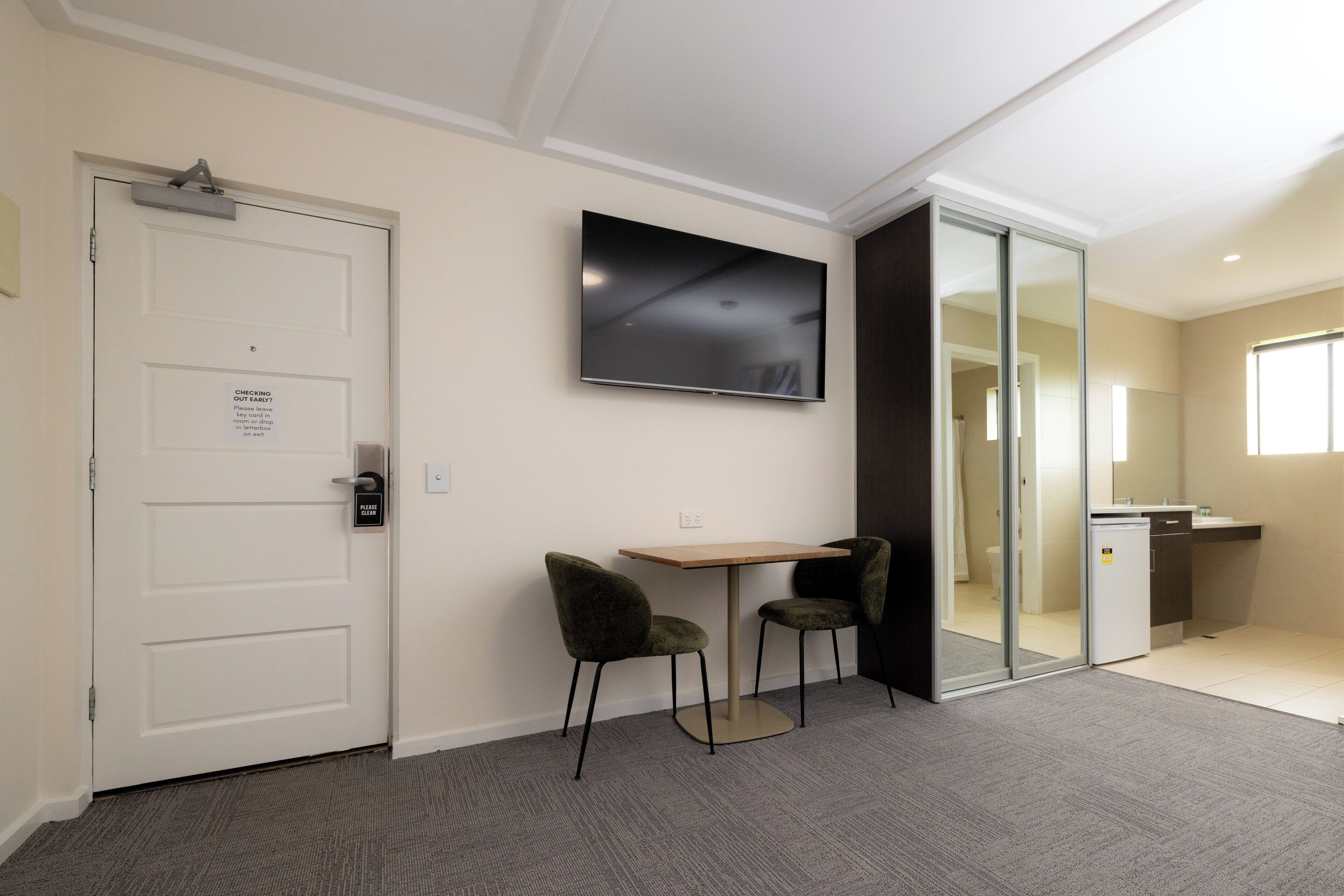 Une chambre d’hôtel avec une porte blanche, un téléviseur à écran plat fixé au mur, une petite table avec deux chaises et une porte-fenêtre coulissante donnant sur une salle de bain.