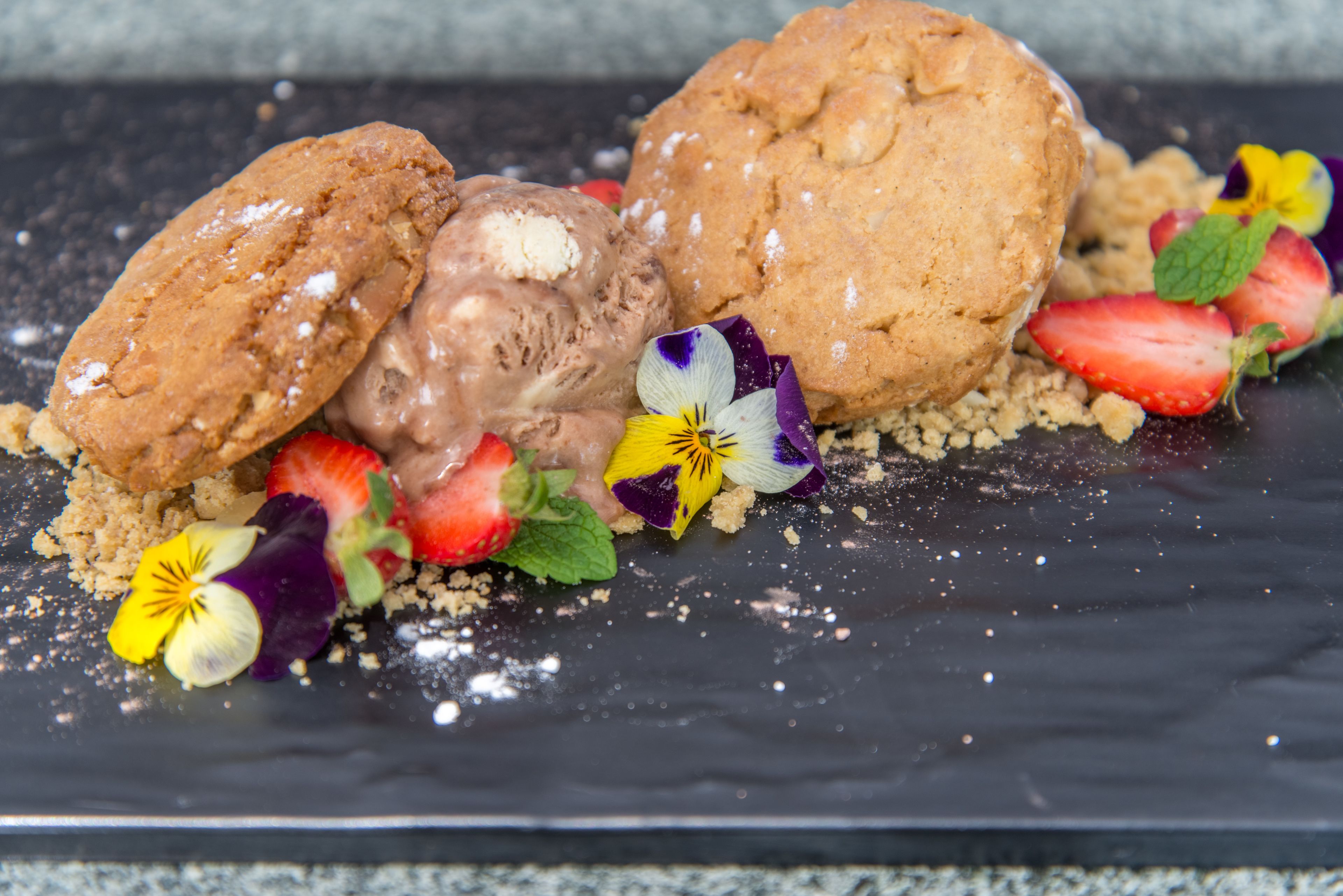 Un postre con galletas, helado, fresas y flores sobre un plato oscuro.