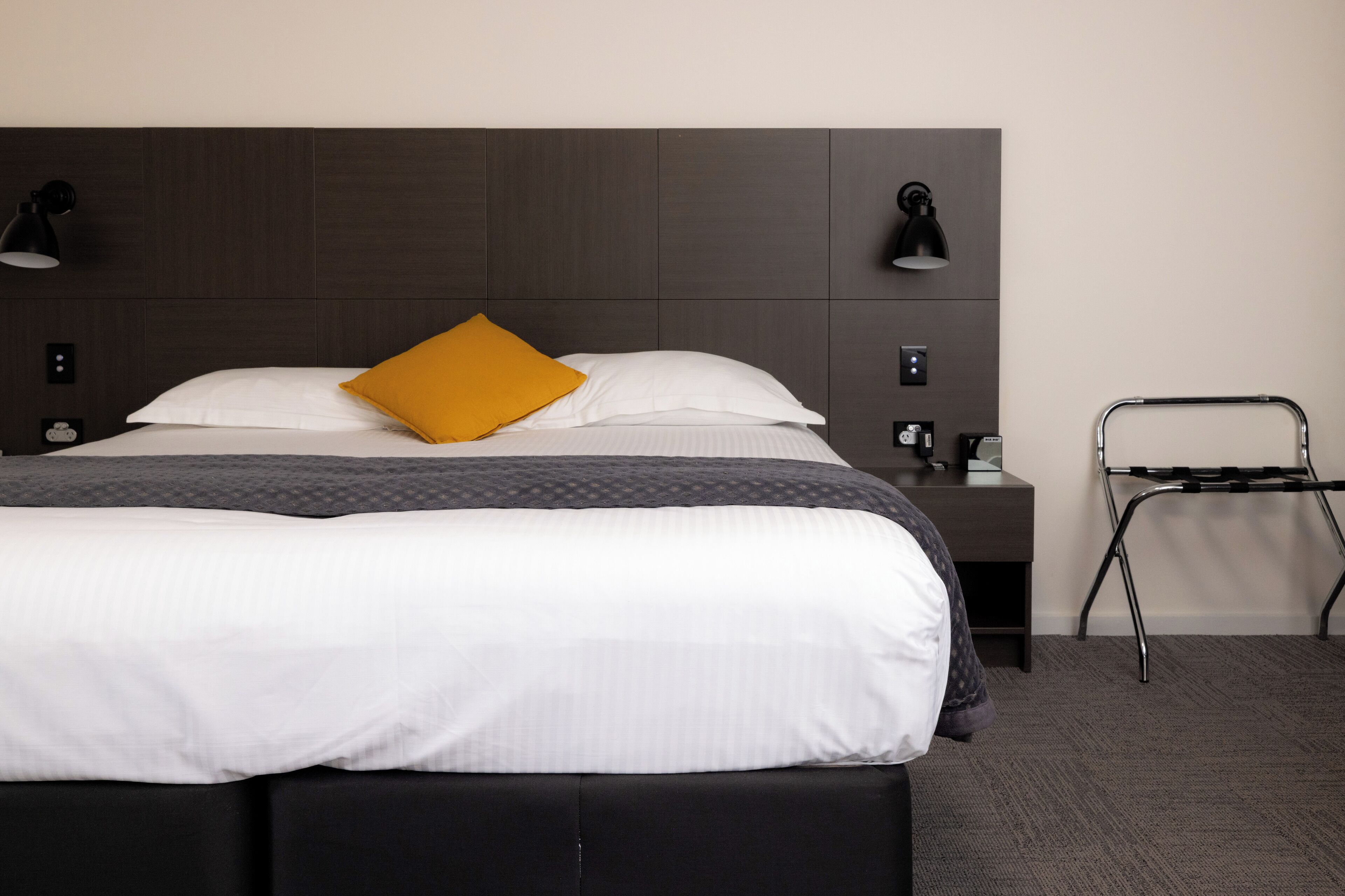 A neatly made bed with white and gray bedding, a mustard yellow pillow, and two black wall-mounted lamps.