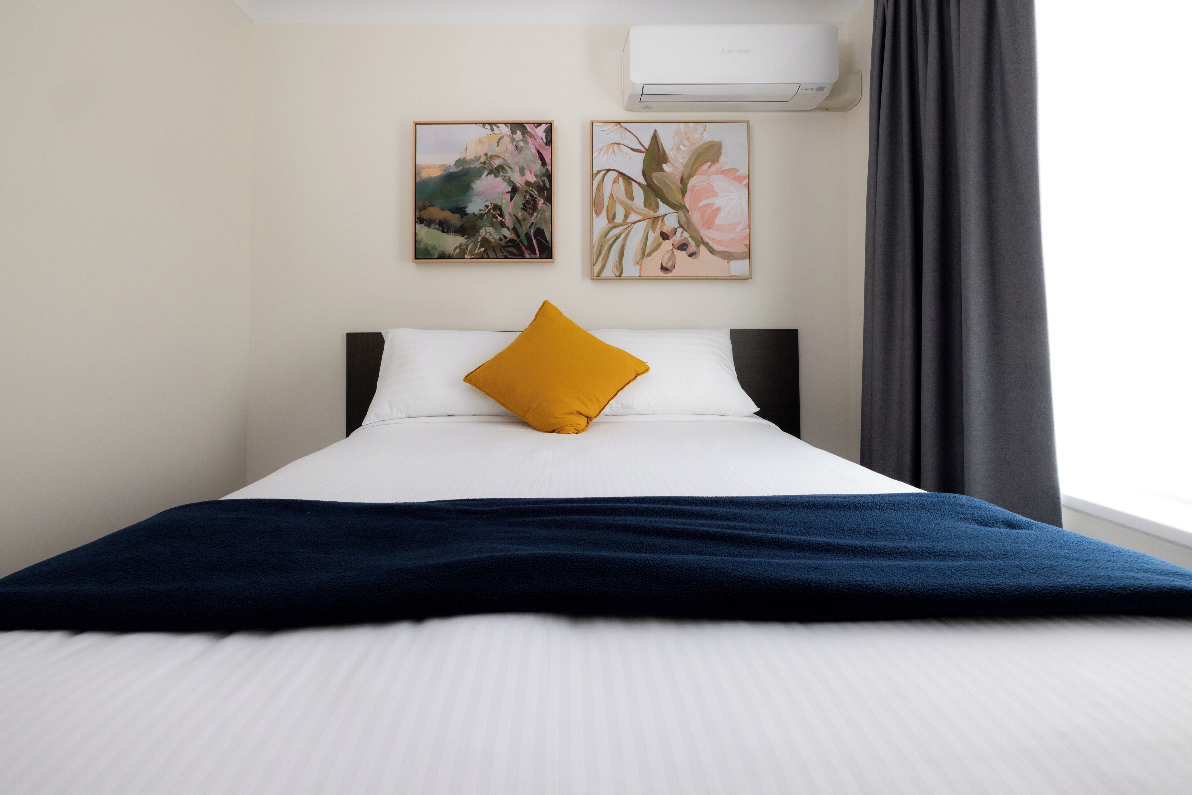 A neatly made bed with a blue blanket, a mustard-colored pillow, and two floral paintings on the wall.
