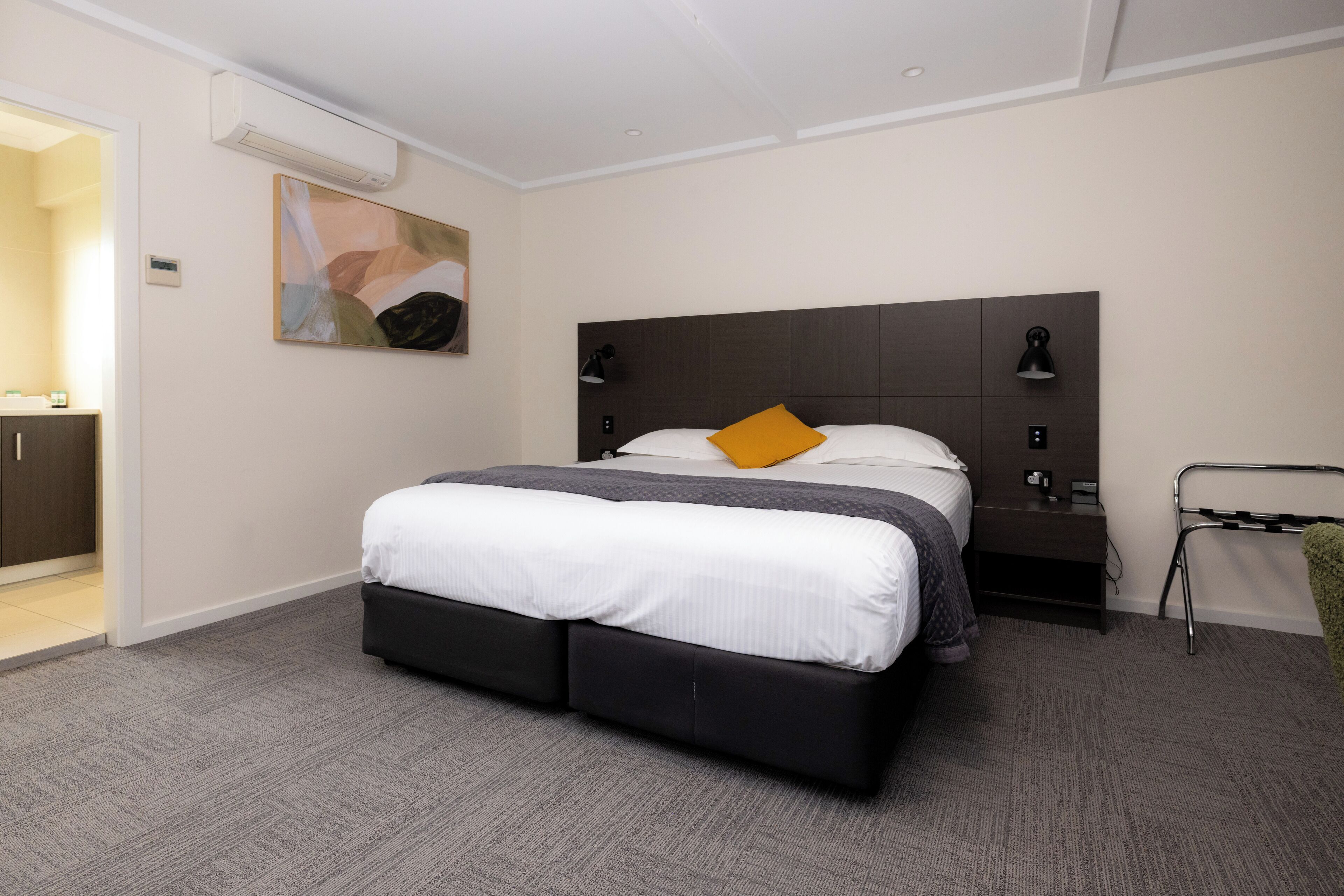 A modern hotel room with a large bed, a bedside table, a wall-mounted air conditioner, and a painting on the wall.