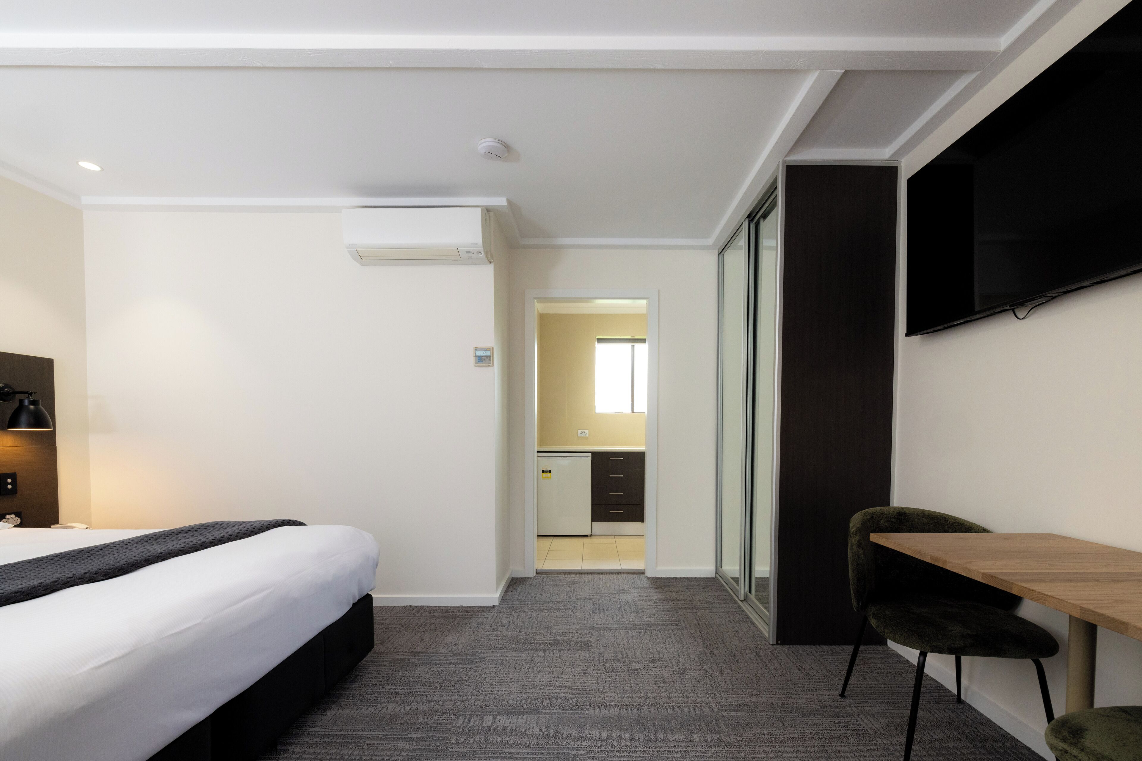 A hotel room with a bed, a desk, a chair, a television, and a kitchenette in the background.