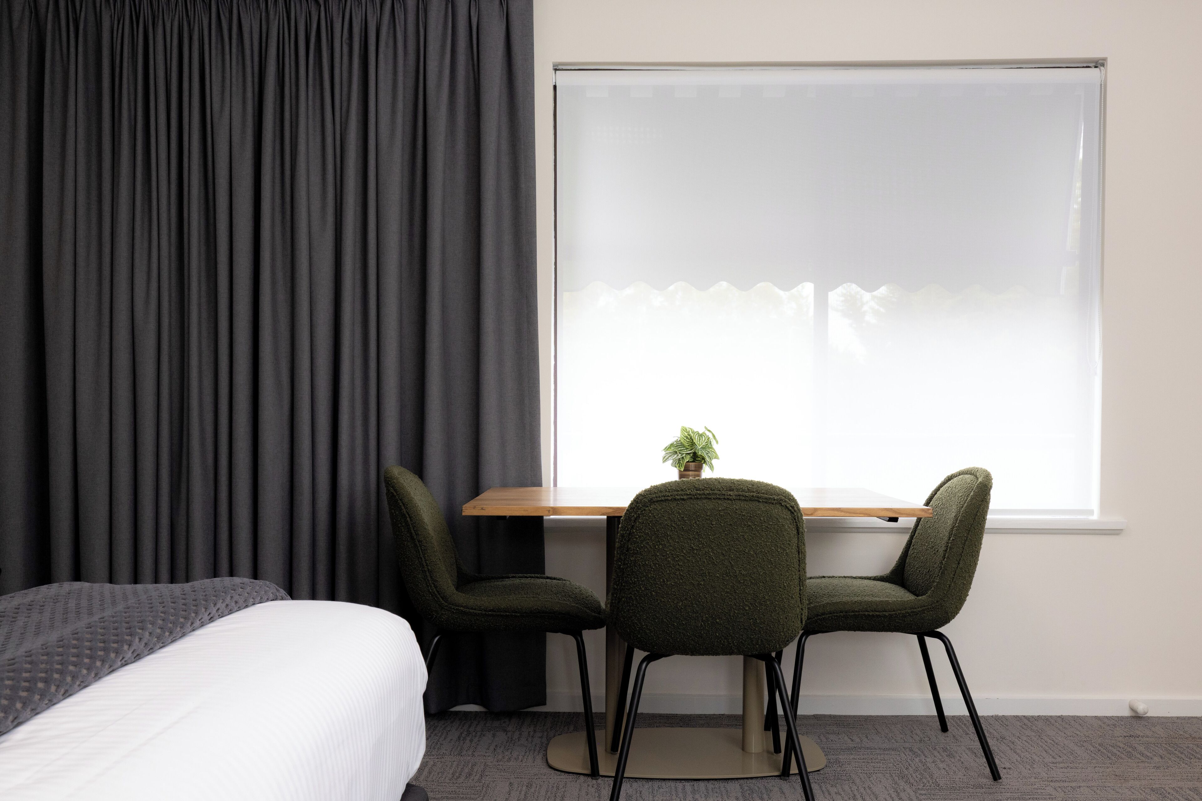 A hotel room with a bed, a desk, two chairs, a potted plant, and a large window with curtains.