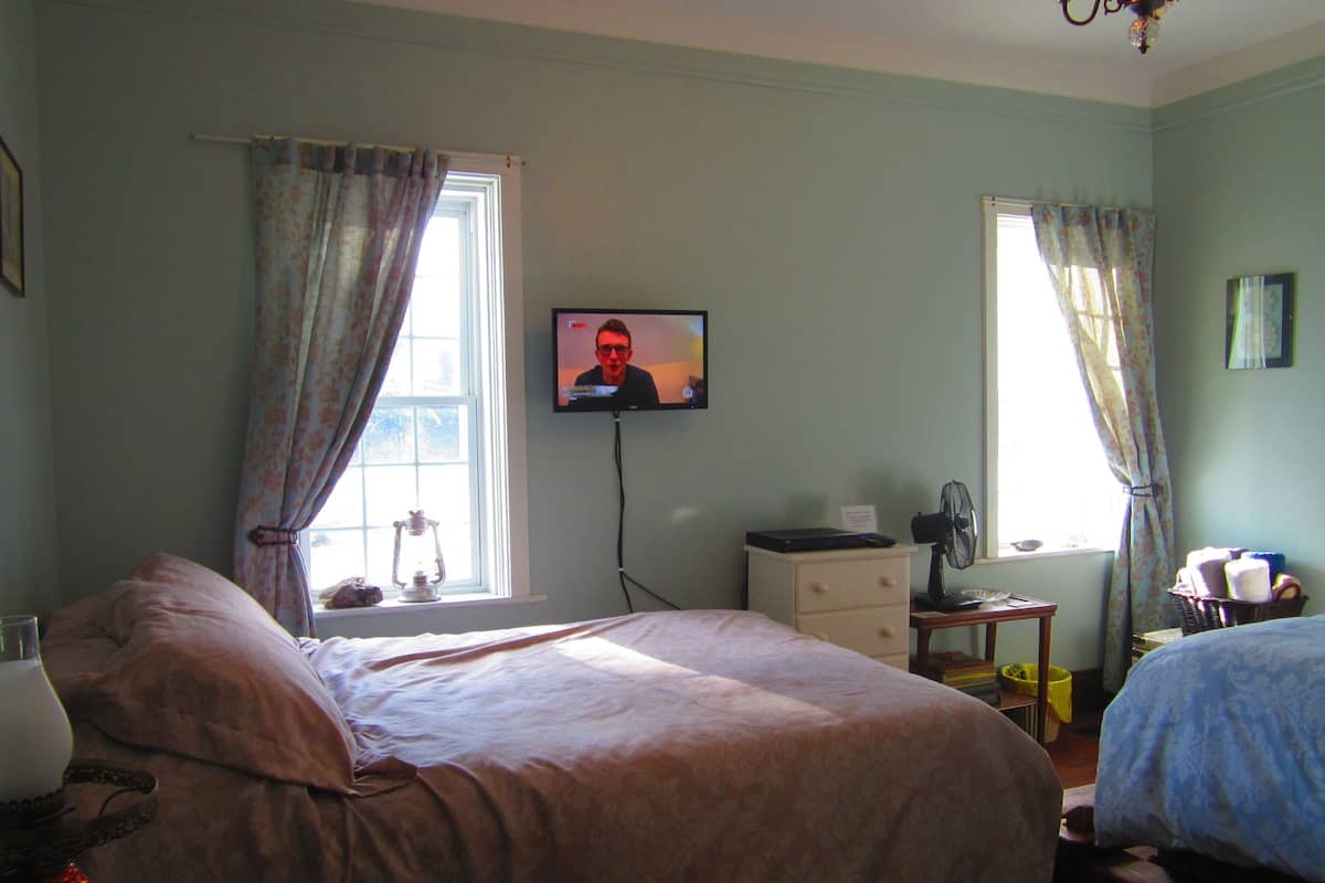 Un dormitorio con cama, televisor en la pared, ventana con cortinas, mesita de noche y lámpara.