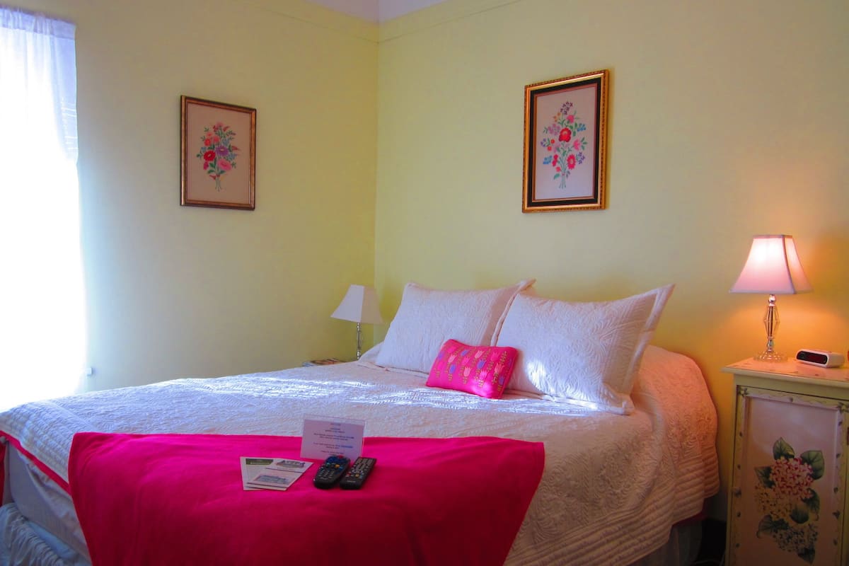 Una cama bien hecha con una manta roja, dos almohadas y una mesita de noche con una lámpara y un reloj. En la pared hay cuadros con motivos florales y una ventana con cortinas.