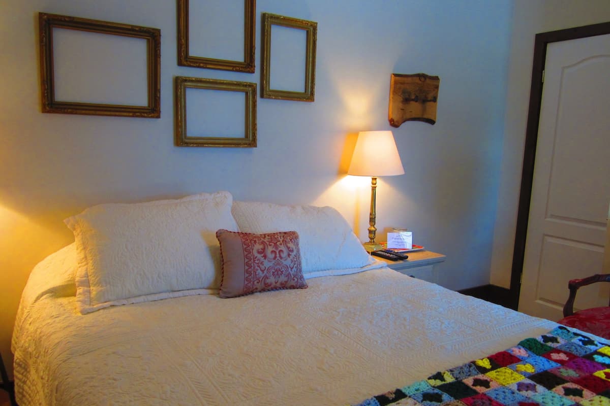 Una cama bien tendida con sábanas blancas y un edredón colorido, una lámpara de noche y una pared con cuadros.