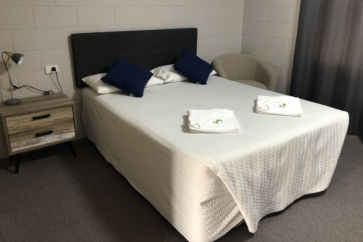 A neatly made bed with pillows, a bedside table with a lamp, and a chair.