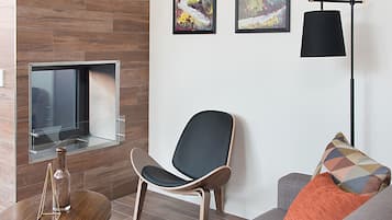 A modern living room with a flat-screen TV mounted on a wooden wall, a black chair, a gray sofa with an orange pillow, and a floor lamp.