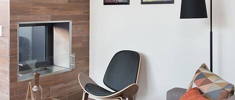 A modern living room with a flat-screen TV mounted on a wooden wall, a black chair, a gray sofa with an orange pillow, and a floor lamp.