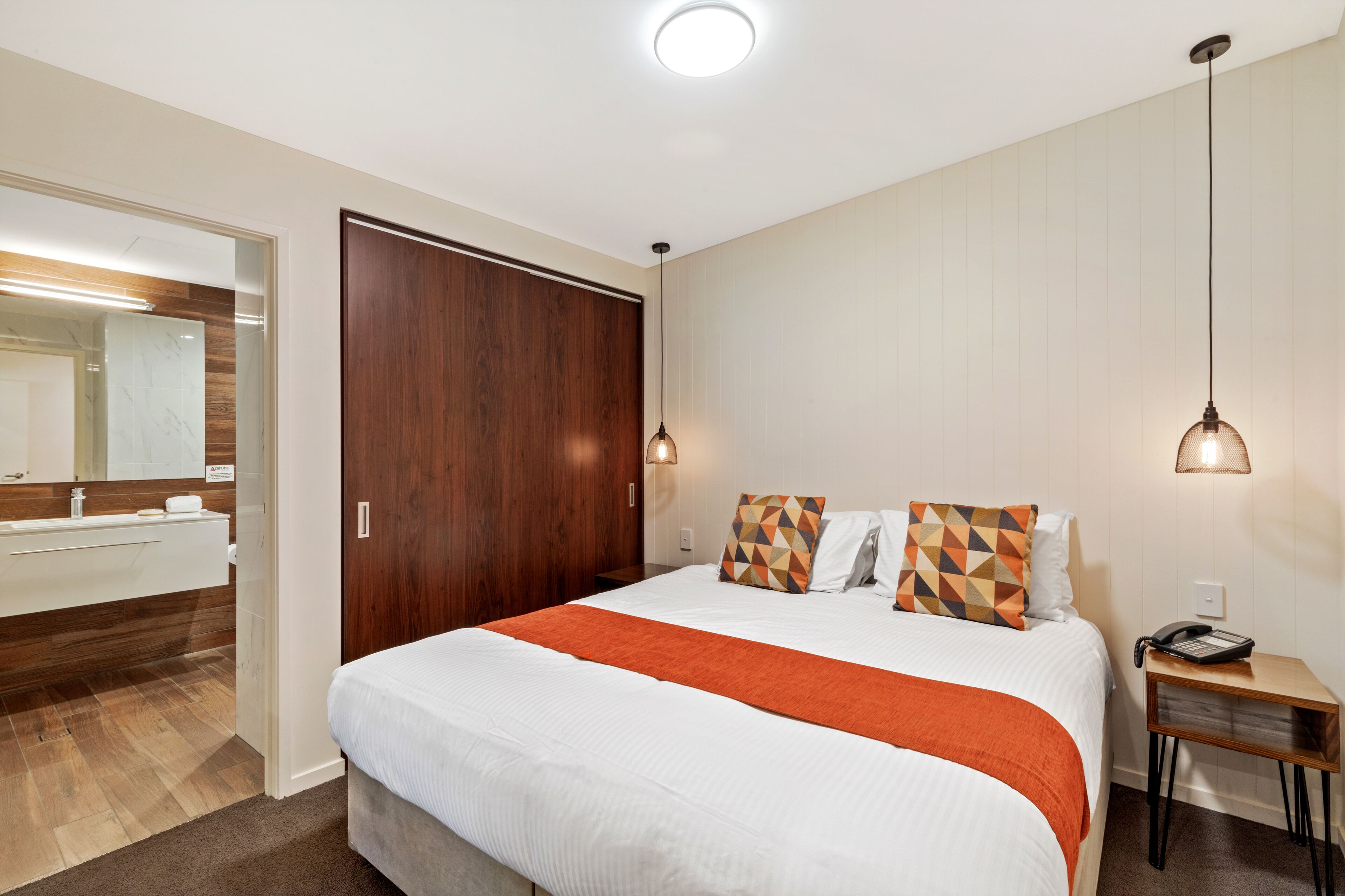 Une chambre d’hôtel avec un grand lit, une armoire en bois, une table de chevet avec une lampe, et une salle de bain visible à travers une porte ouverte.