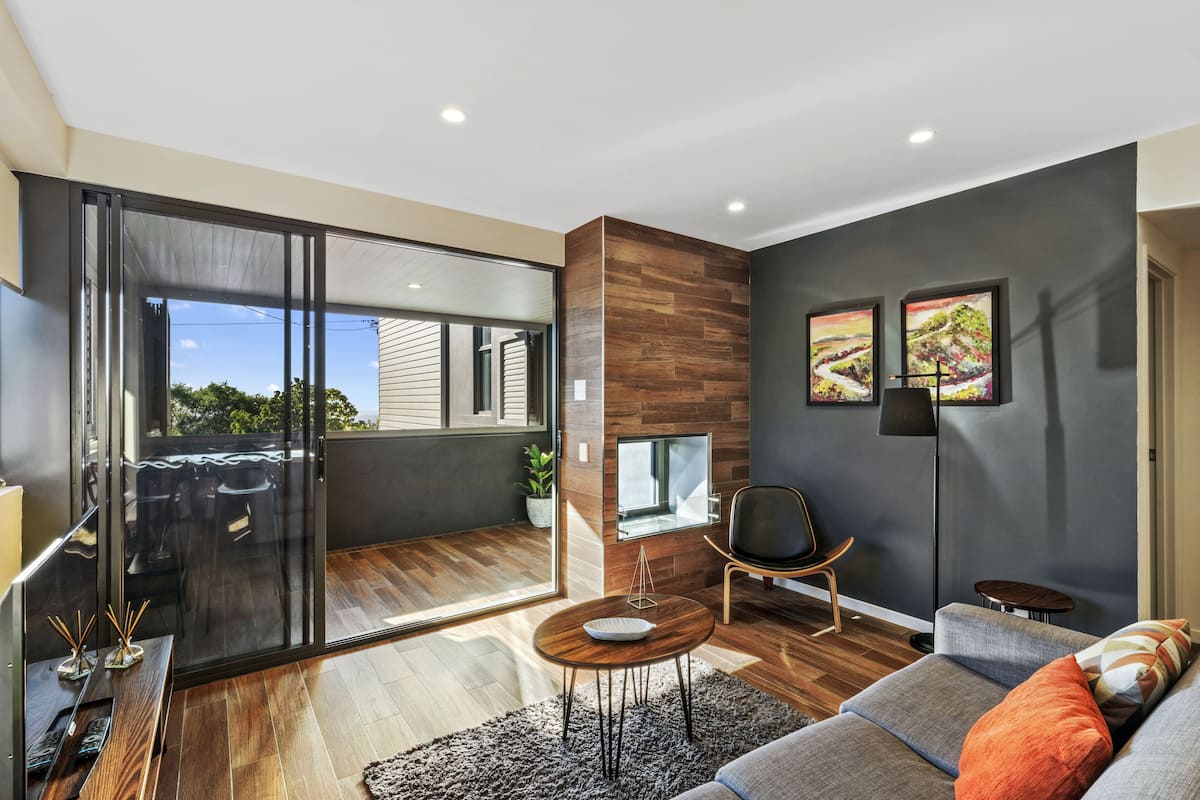 A modern living room with a sliding glass door leading to a balcony, a grey sofa with orange cushions, a wooden coffee table, and a black floor lamp.