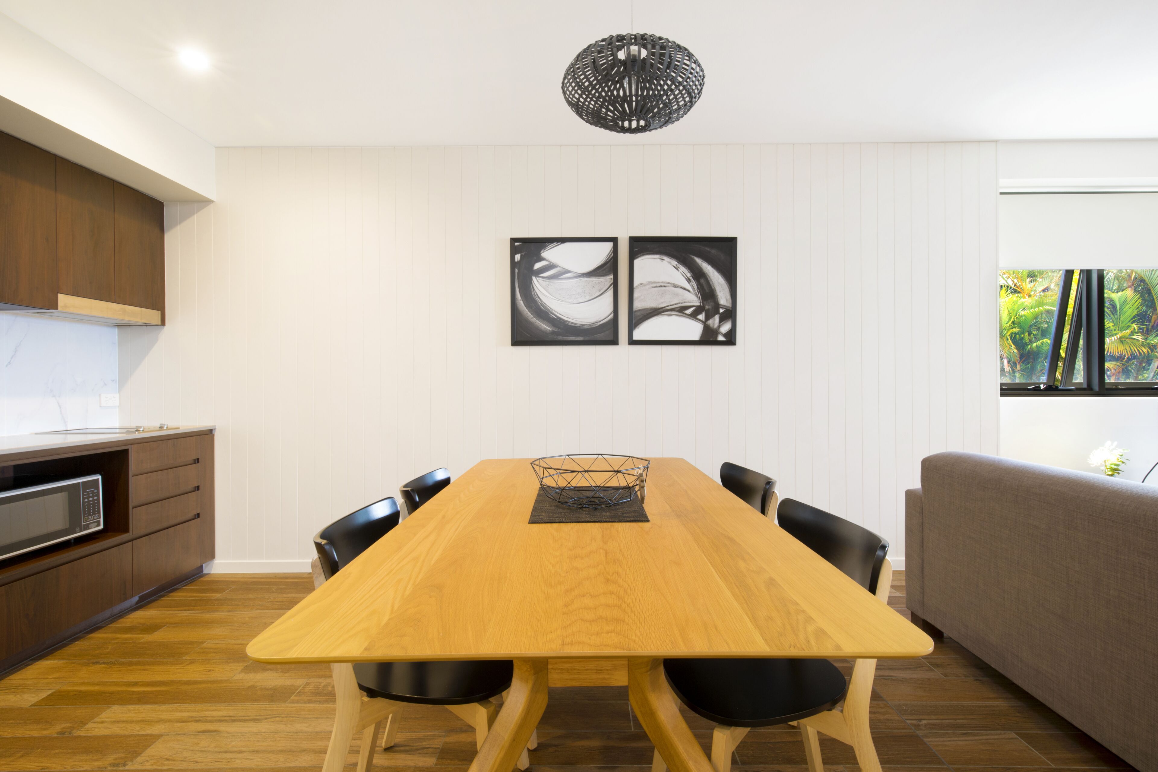 A modern kitchen with wooden cabinets, a microwave, and a dining area with a wooden table and chairs. There are two black and white artwork pieces on the wall, and a window with a view of greenery.