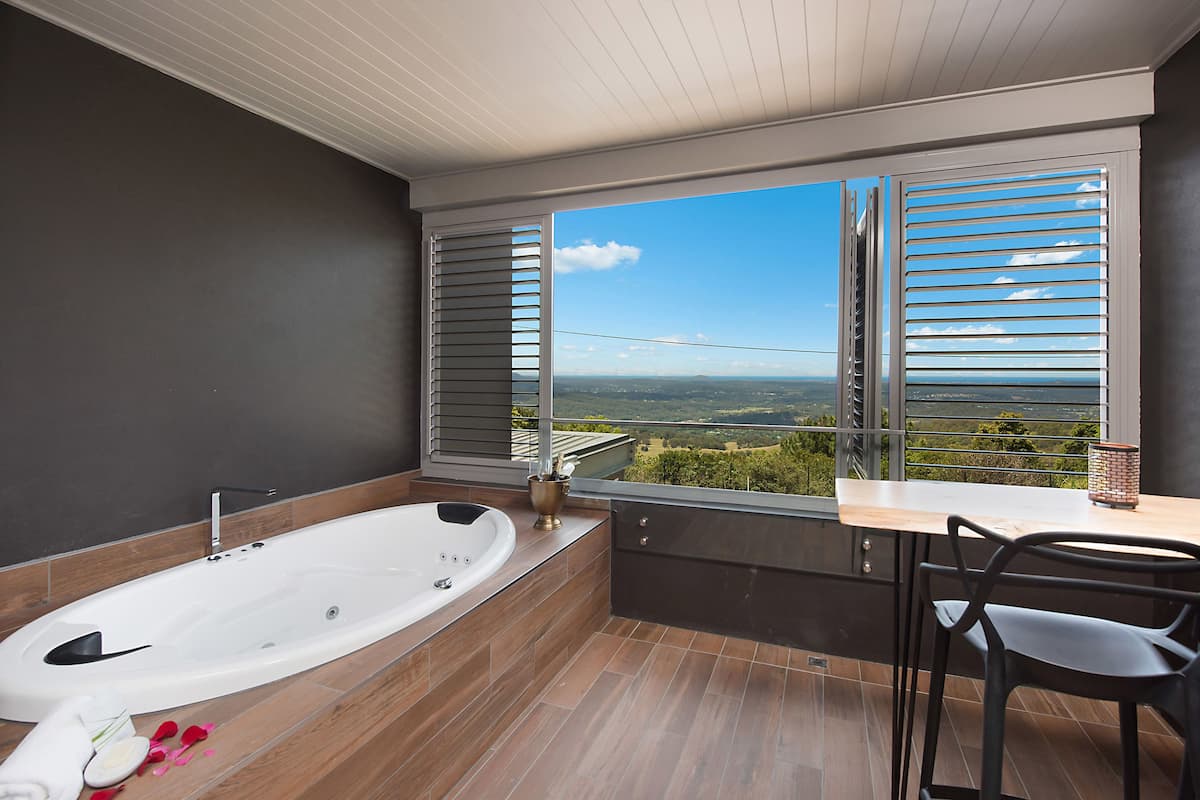 A modern bathroom with a large bathtub, a shower, and a view of the countryside through a window with blinds.