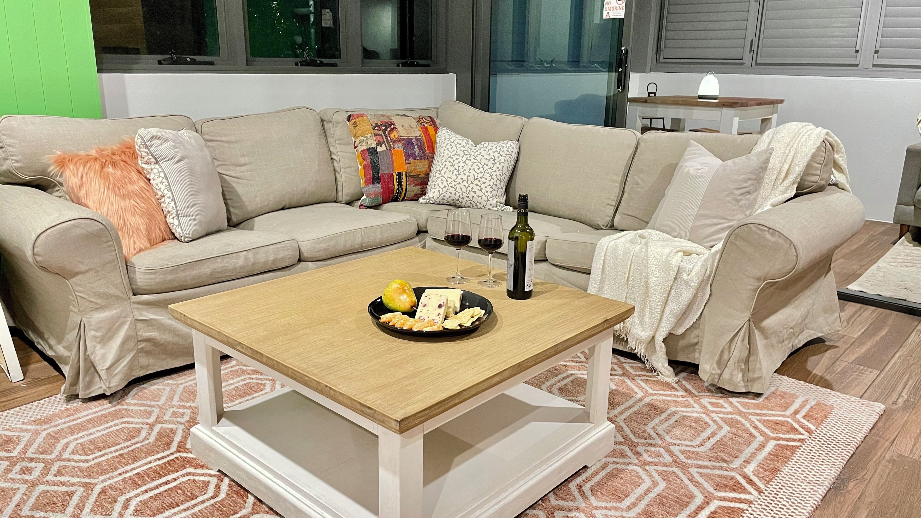 A living room with a beige sectional sofa, a wooden coffee table with a plate of snacks and a bottle of wine, and a patterned rug.
