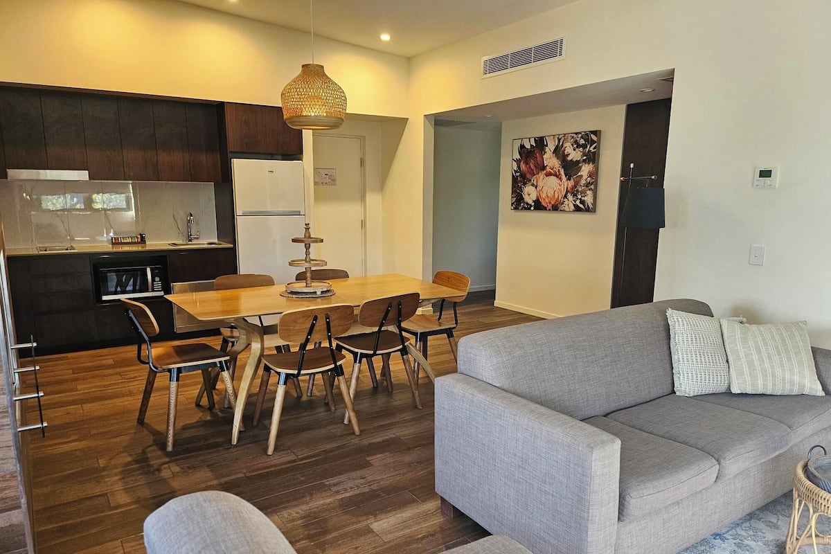 A modern living room with a grey sofa, a dining table with chairs, a kitchen area with a refrigerator and microwave, and a hanging light fixture.
