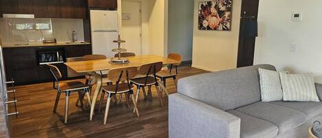 A modern living room with a grey sofa, a dining table with chairs, a kitchen area with a refrigerator and microwave, and a hanging light fixture.