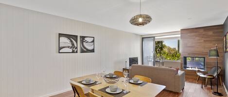Ein moderner Essbereich mit Holztisch, Stühlen und einer Couch. An der Wand befinden sich abstrakte Gemälde und ein großes Fenster mit Blick auf Grünflächen.