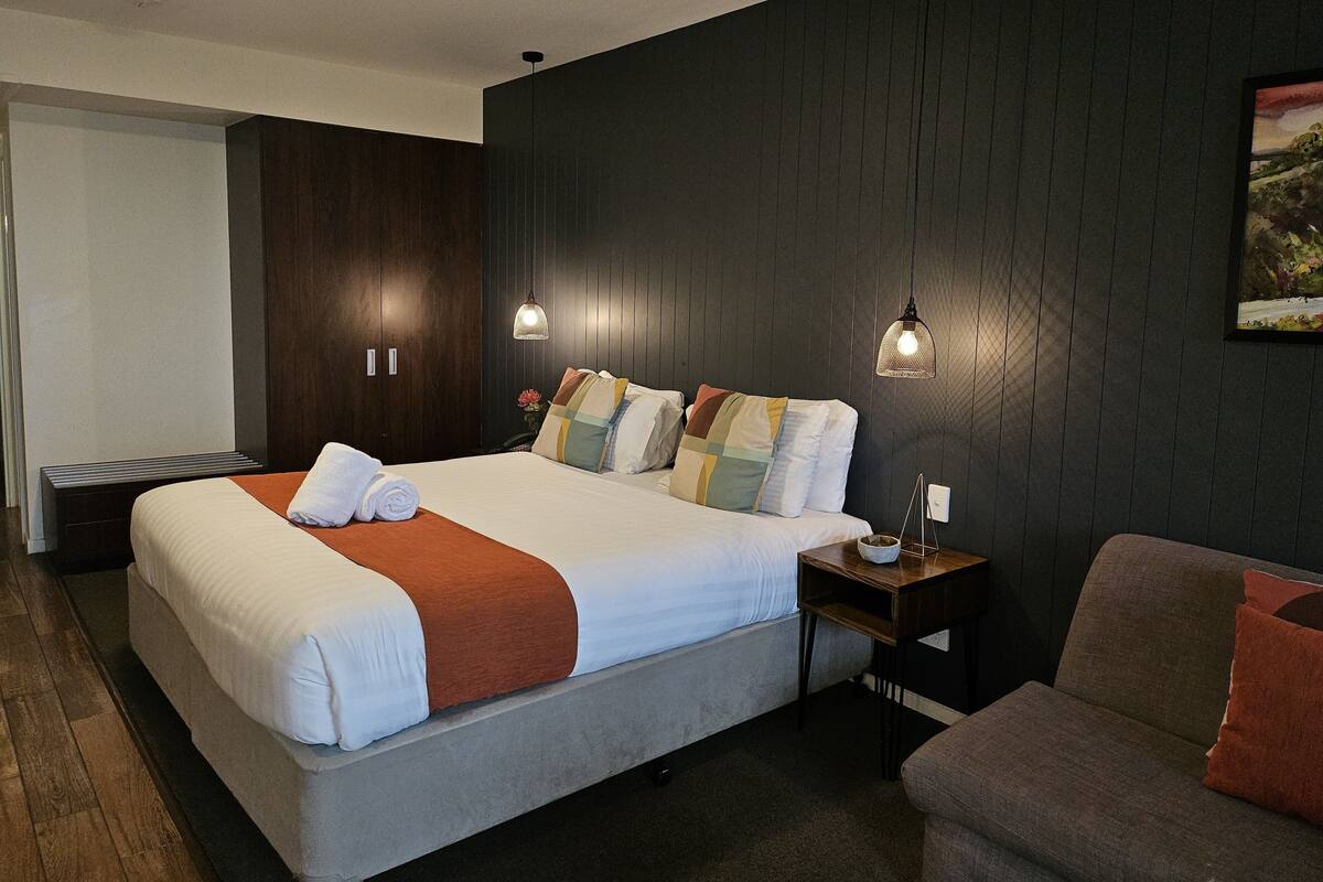 A modern hotel room with a large bed, a bedside table, a chair, and a dark wooden wall.
