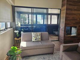 Una sala de estar con sofá, una mesa de centro y televisor montado en una pared de madera. Hay una puerta corrediza de vidrio que da a un área exterior con sillas.