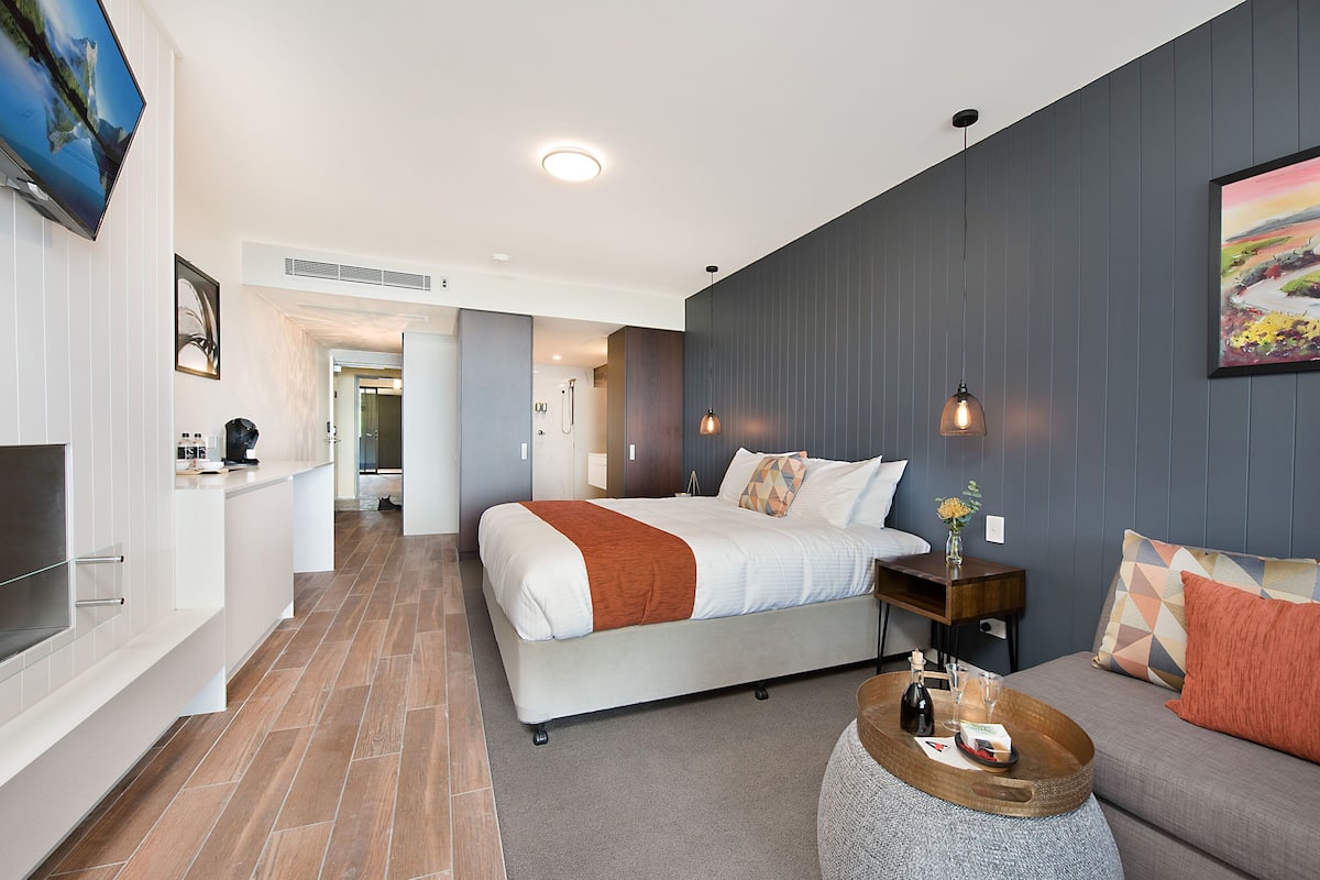 A modern hotel room with a large bed, a sofa, a TV, and a dining area.