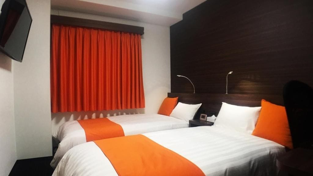 A hotel room with two beds, a television mounted on the wall, and a window with red curtains.