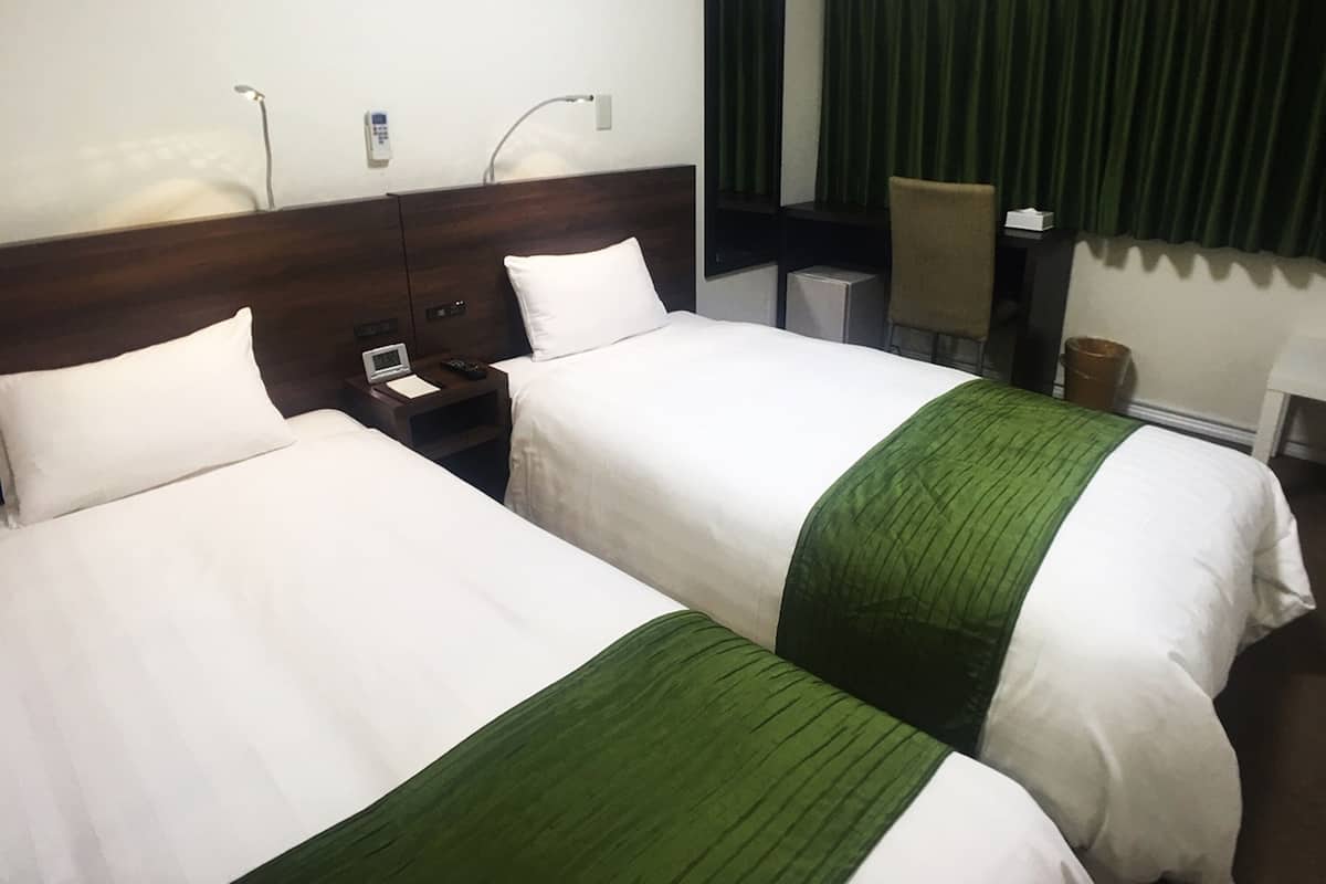 Two adjacent hotel beds with white linens and green accents, a bedside table with a phone and a lamp, and a green curtain in the background.