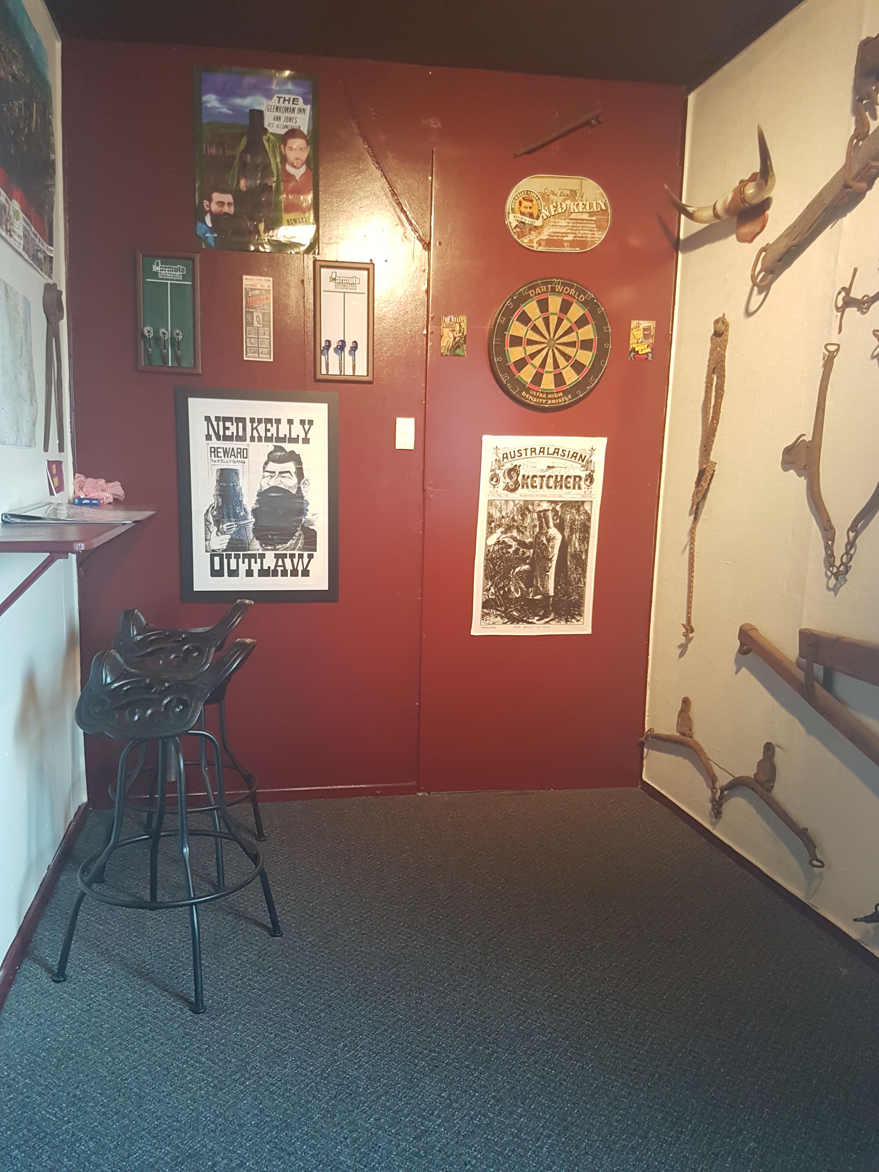 A room with a red wall, a dartboard, a framed poster of Ned Kelly, and a metal cat-shaped stool.