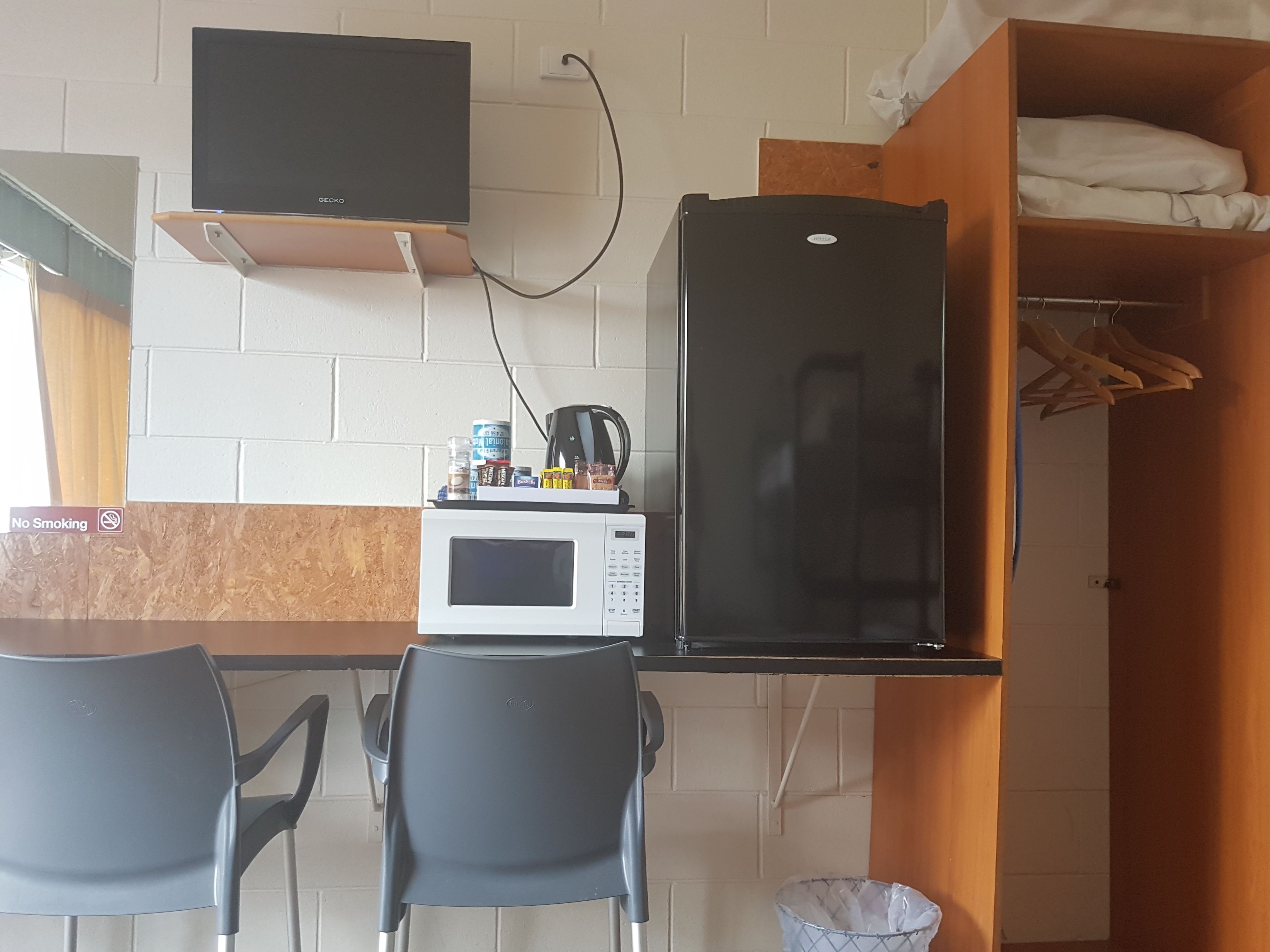 A compact kitchenette with a microwave, a black refrigerator, and a small countertop with various items. There are two chairs and a trash bin.
