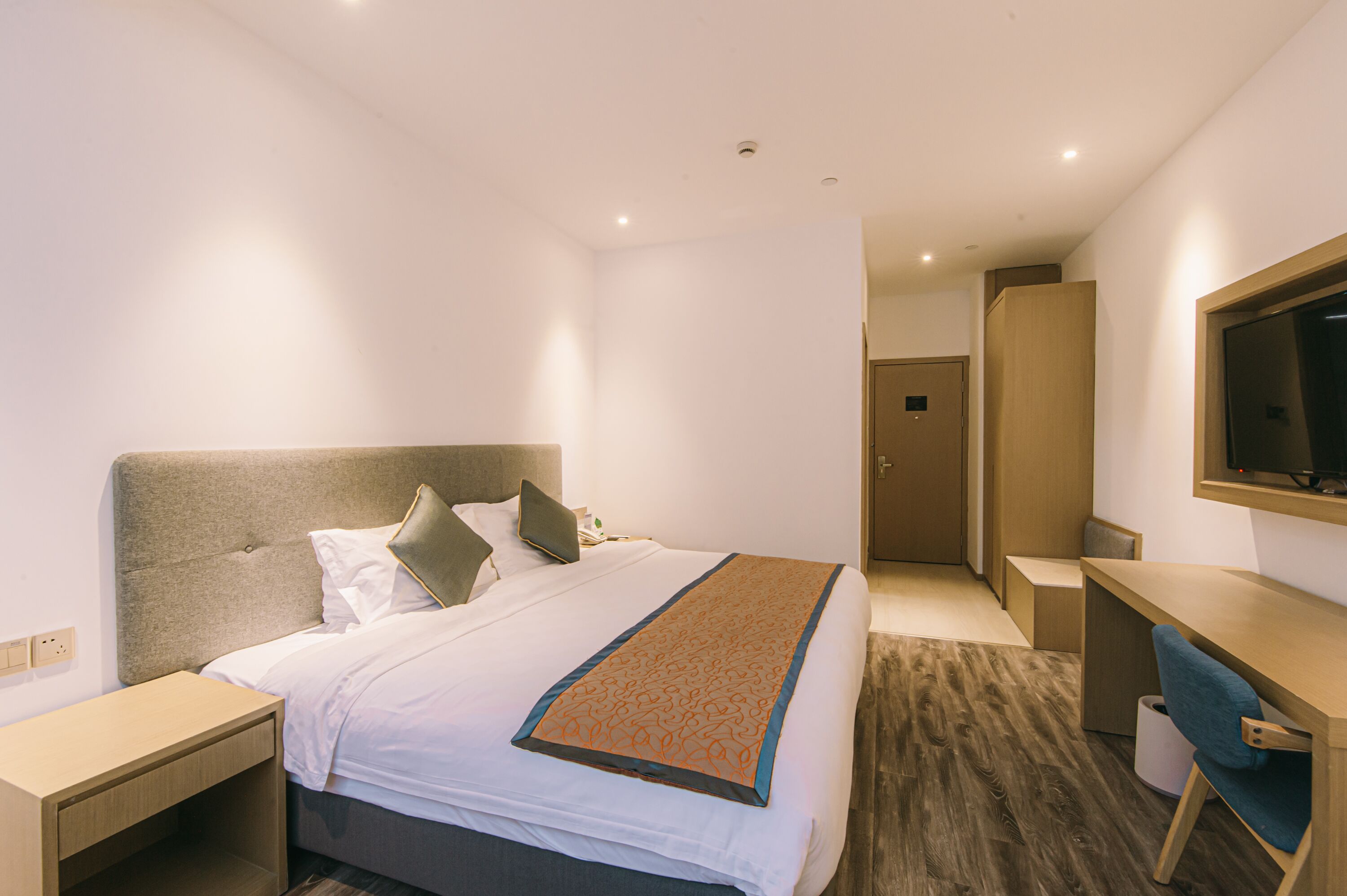 A hotel room with a bed, bedside table, desk, chair, and a TV mounted on the wall.