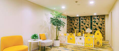A brightly lit room with a yellow chair, a small round table, a potted plant, and a play area with yellow and white toys.
