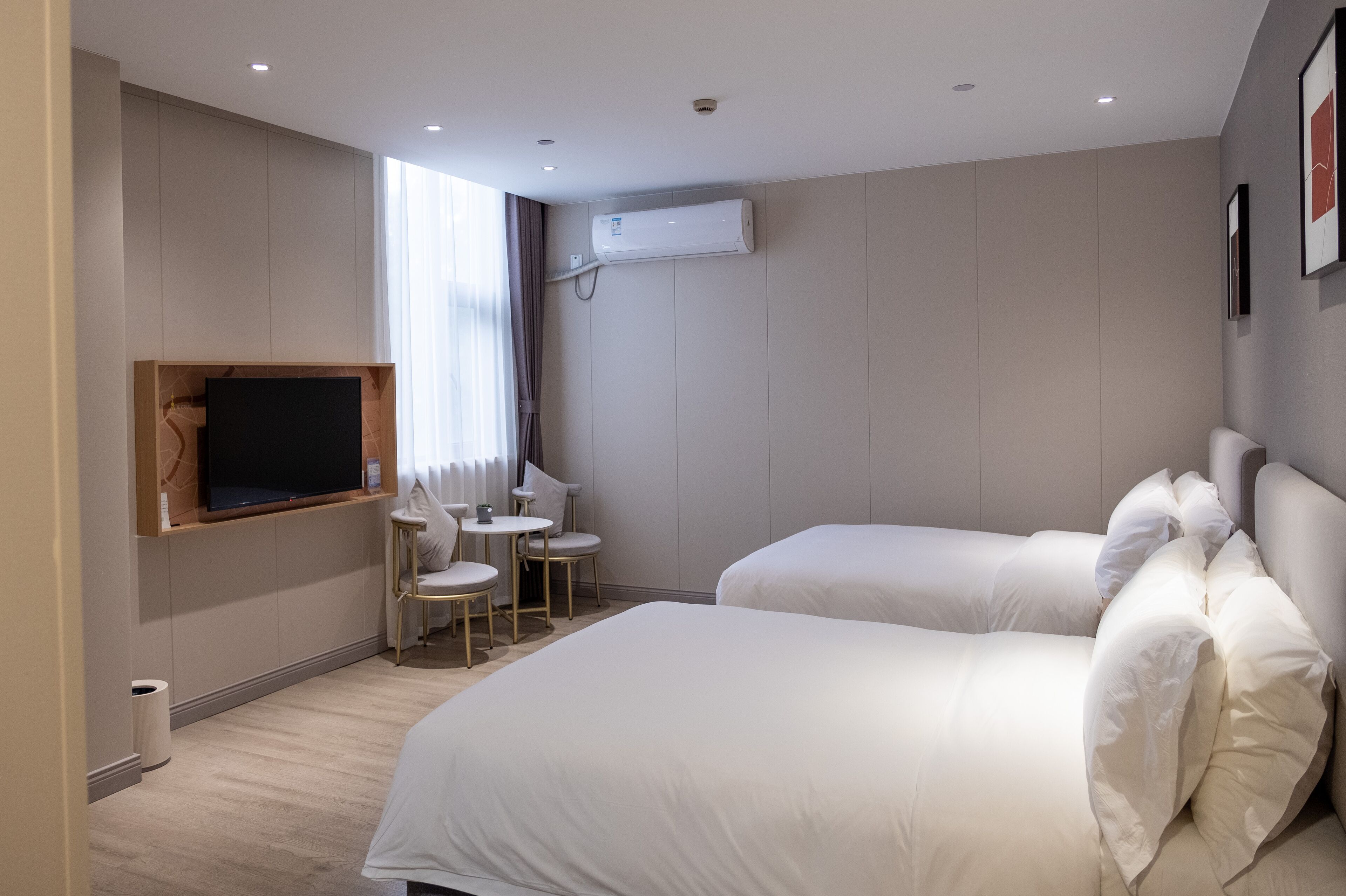 Hotellihuone, jossa on kaksi sänkyä, seinälle asennettu televisio, pieni pöytä tuoleineen ja ilmastointilaite.