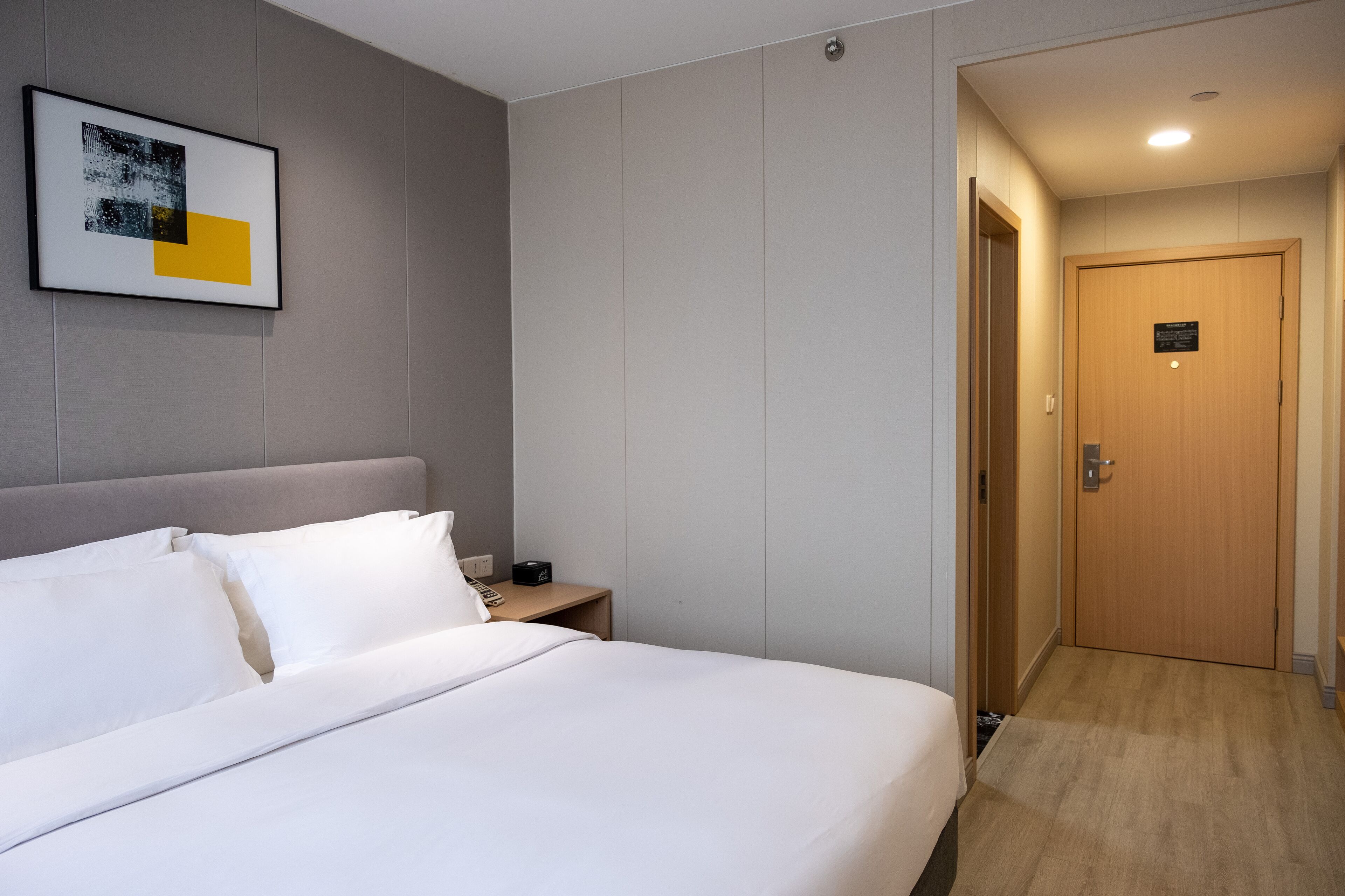 A hotel room with a bed, a nightstand, a framed picture on the wall, and a wooden door.