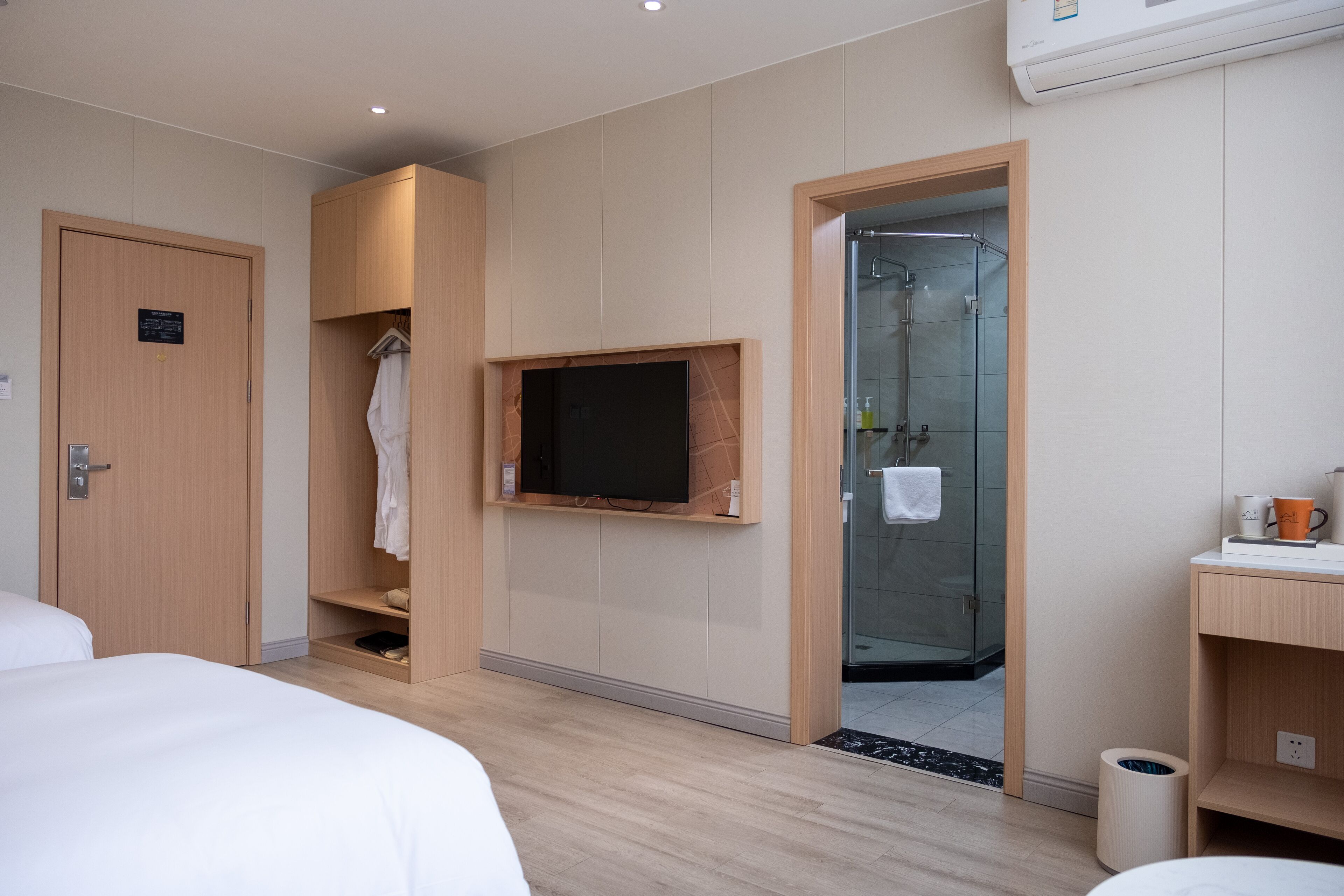 A hotel room with a bed, a TV mounted on the wall, a shower area, and a small shelf with a cup and a bottle.