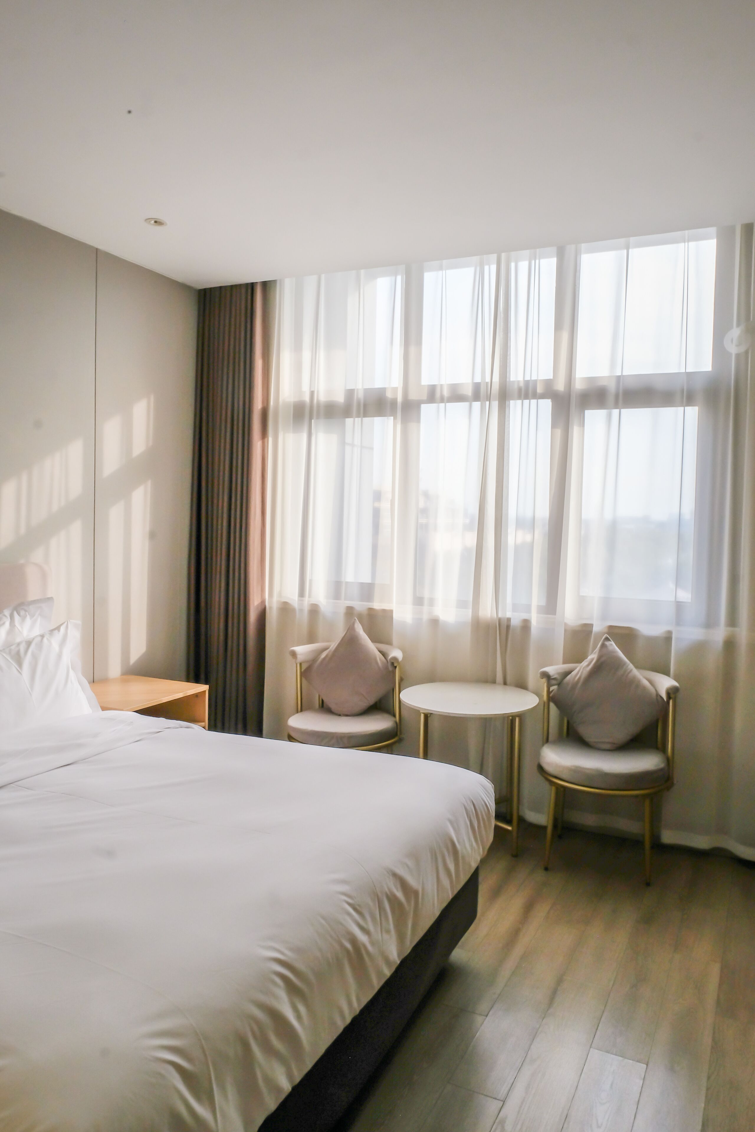 A hotel room with a large bed, two armchairs, a small table, and a window with sheer curtains.