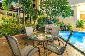 A round table with chairs and a poolside setting with a grill and plants. - Butterfly Garden - A Boutique Residence (Pattaya)
