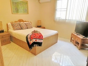 A bedroom with a large bed, a television on a wooden stand, and a window with blinds.