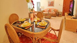 A dining area with a round table set for four, surrounded by wicker chairs with red cushions. A television is visible in the background, and a sofa is placed in the living area.