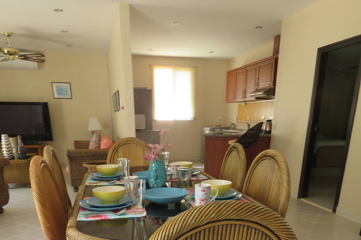 A dining area with a round table set for six, surrounded by wicker chairs with pink cushions.