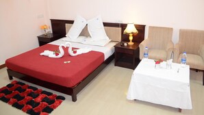 A hotel room with a bed, a bedside table with a lamp, a chair, and a small table with a vase and water bottles. - KSTDC Hotel Mayura Chalukya Badami (Badami)