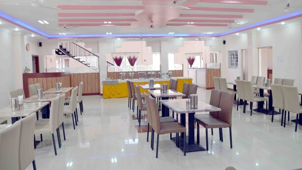 A spacious dining area with multiple tables and chairs, a central counter, and a staircase in the background. - KSTDC Hotel Mayura Chalukya Badami (Badami)