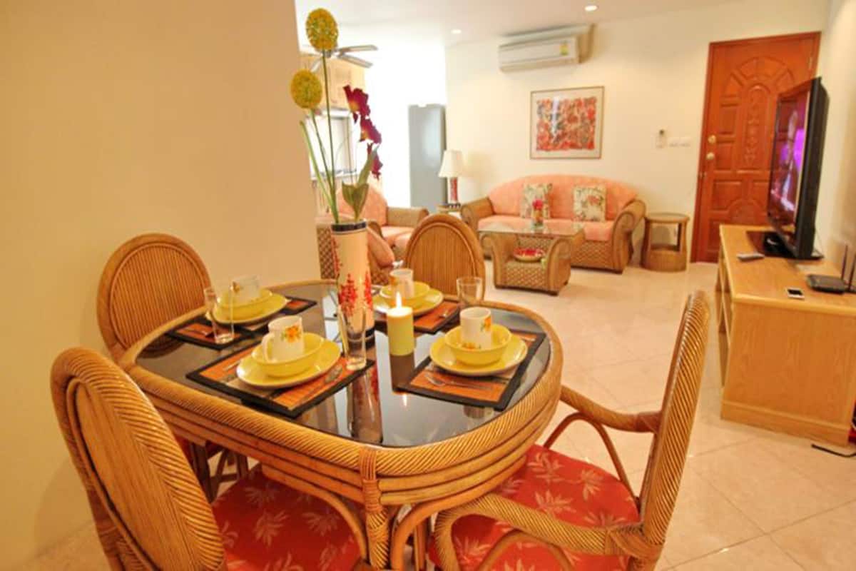 Un coin repas avec une table ronde dressée pour quatre personnes, entourée de chaises en osier avec des coussins rouges. On aperçoit un téléviseur à l’arrière-plan, et un canapé est placé dans l’espace salon.