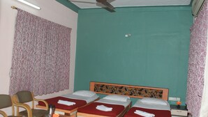 Una habitación con tres camas individuales, un cabecero de madera, una mesita con una jarrón y una flor, y un ventilador de techo.