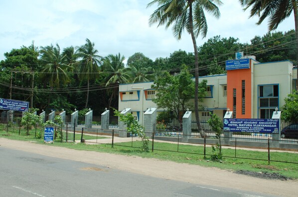 A hotel with a signboard, a fenced area, and palm trees in the background. - KSTDC Hotel Mayura Vijayanagara (Hospet)